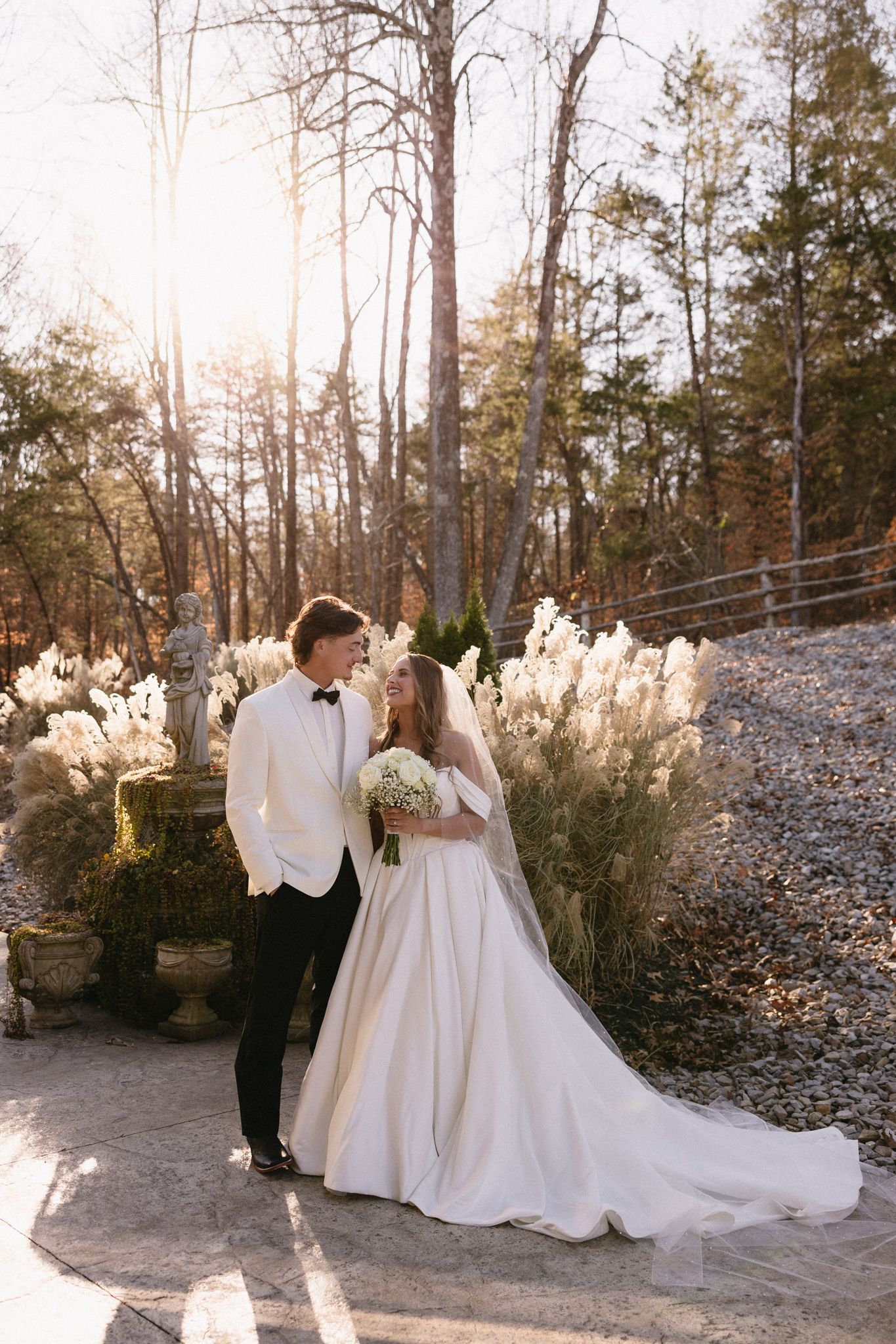 Romantic winter wedding couple poses together in front of tall trees and pampas grass at sunset.
