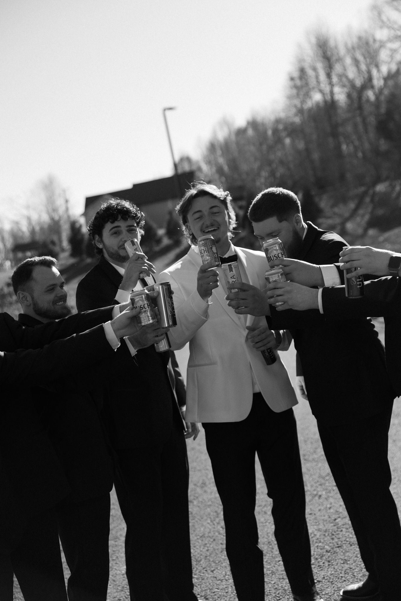 Groomsmen in black suits lined up taking celebratory drinks outdoors.