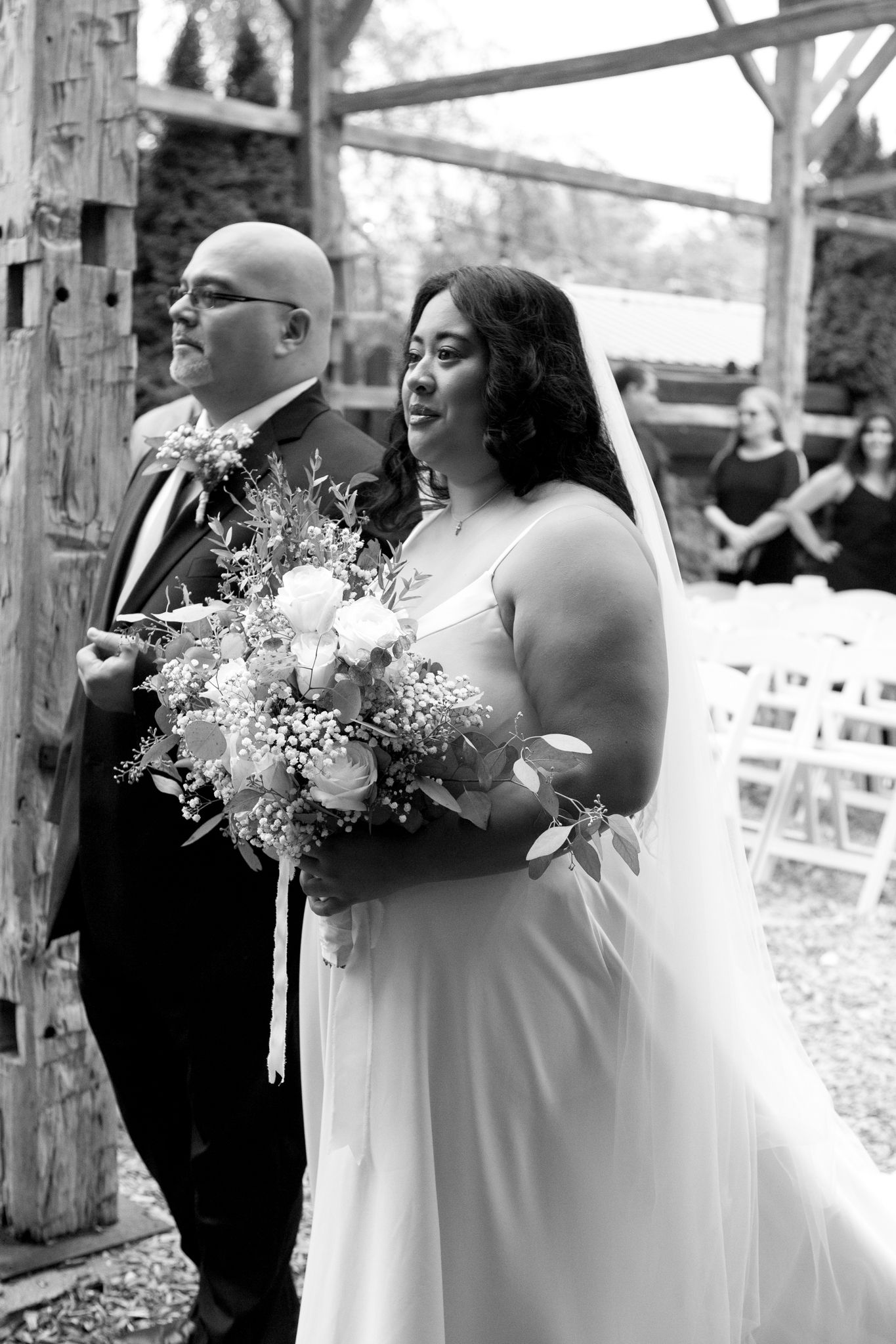 Black and white photo of newlyweds in a rustic outdoor ceremony venue with the bride holding a delicate floral bouquet.