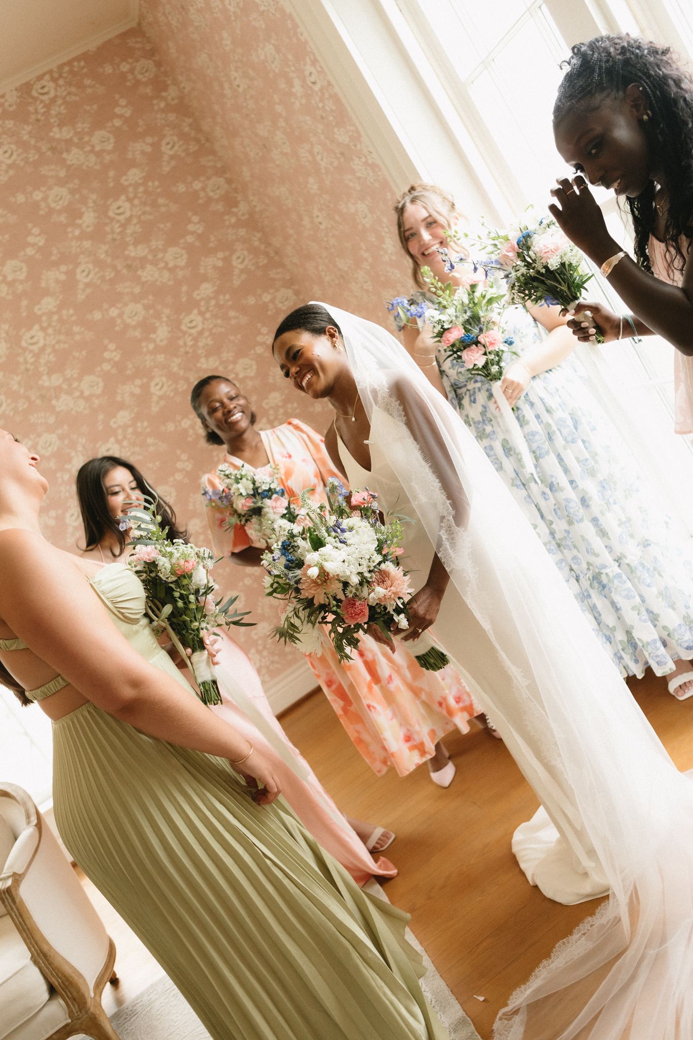 Wedding party poses in olive green and white attire with floral bouquets in a sunlit room.