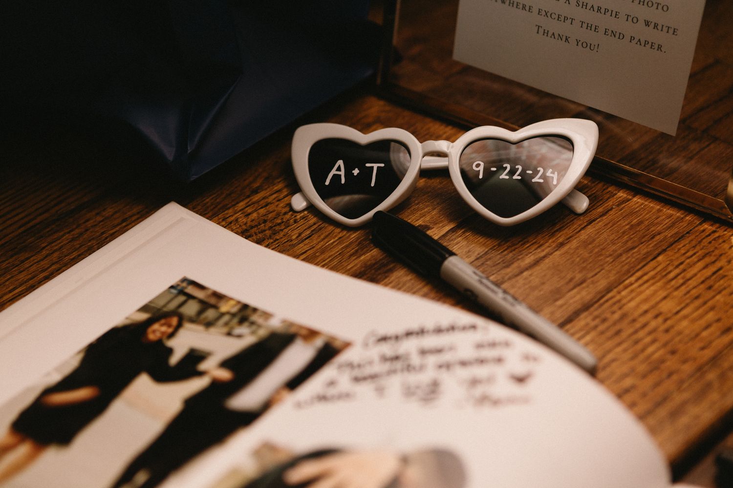 White heart-shaped sunglasses with initials sit atop wedding photos on a rustic wooden surface.
