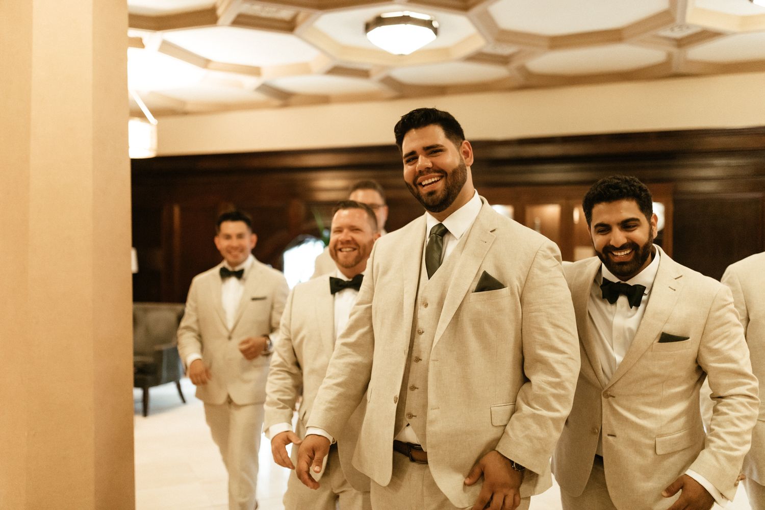 Wedding party members in cream colored suits and black bow ties pose together in an elegant indoor venue.