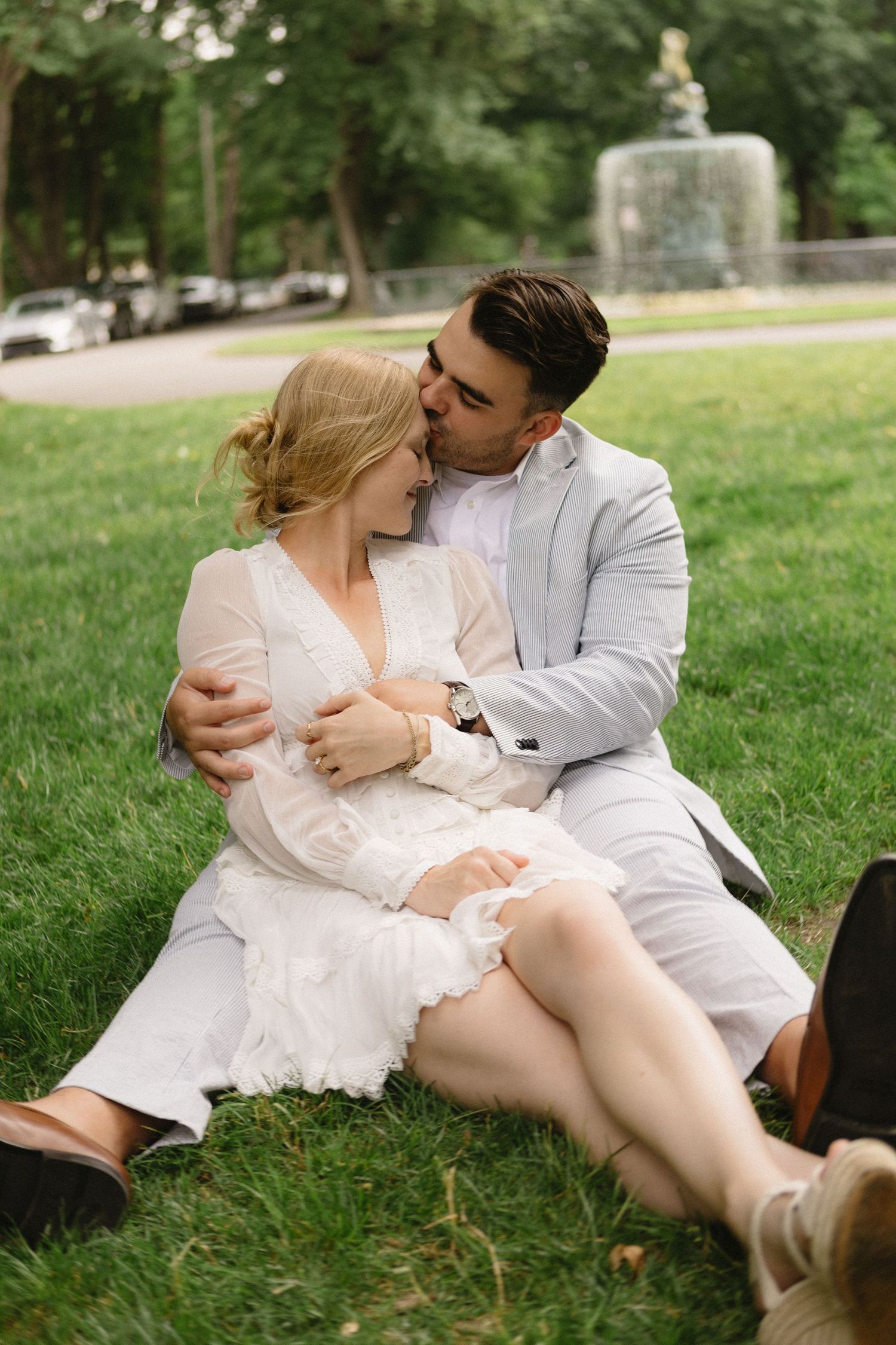 Couple in wedding attire sitting on green grass in a park, sharing a romantic kiss.