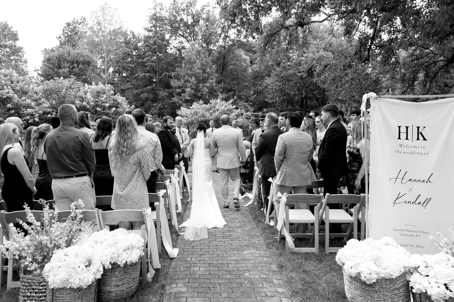 Black and white outdoor wedding ceremony with guests seated along a brick aisle under tall trees.