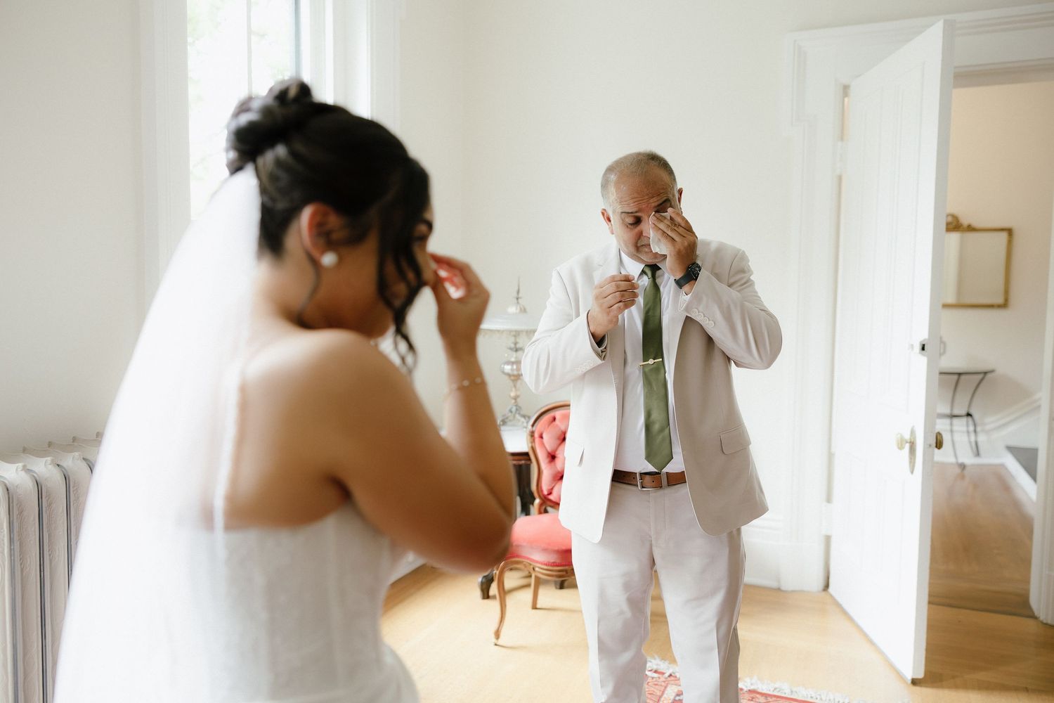 Father of the bride wiping tears while seeing daughter in wedding dress during bridal preparation.