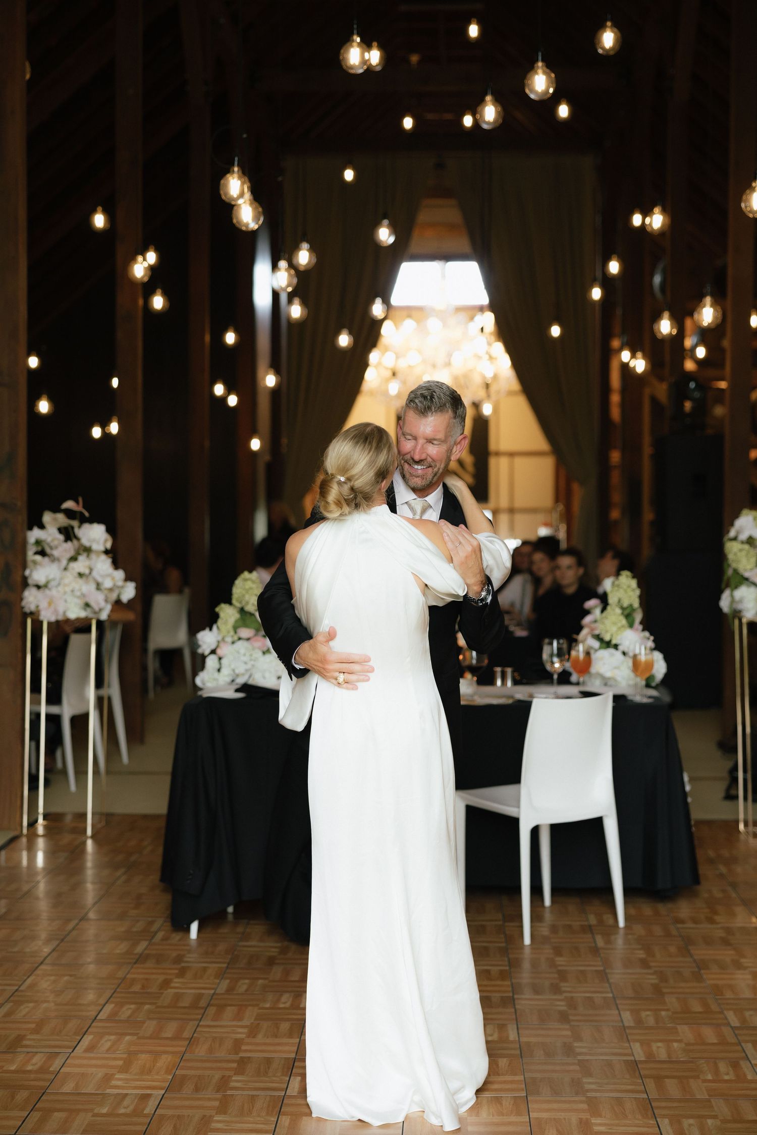 Bride and groom share first dance in rustic barn venue adorned with warm string lights.