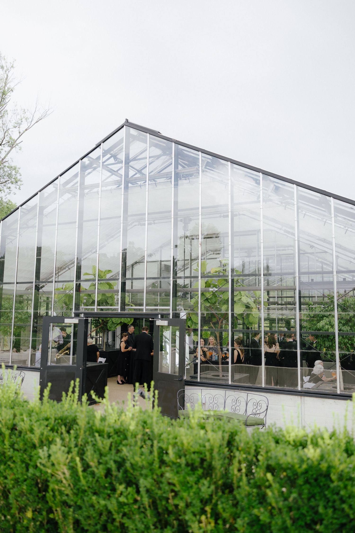 Elegant glass greenhouse wedding venue surrounded by lush greenery with guests mingling inside.