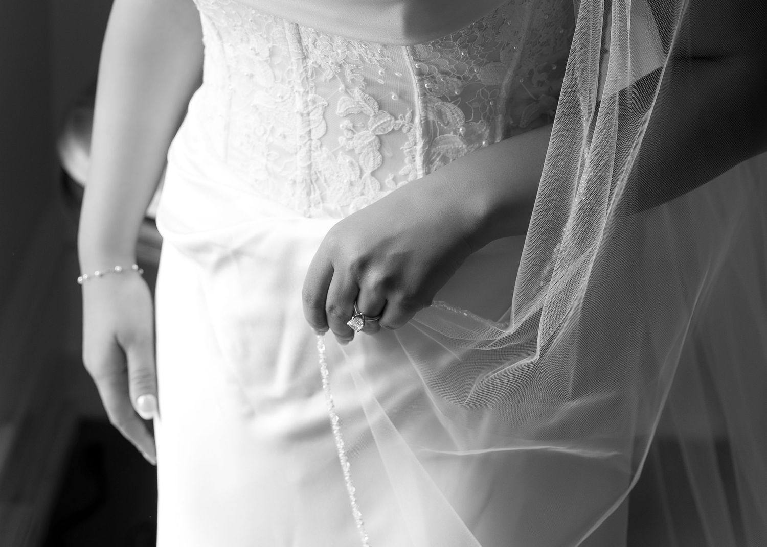 Close-up black and white photo of a bride in a lace wedding dress holding her veil, showcasing an elegant ring.