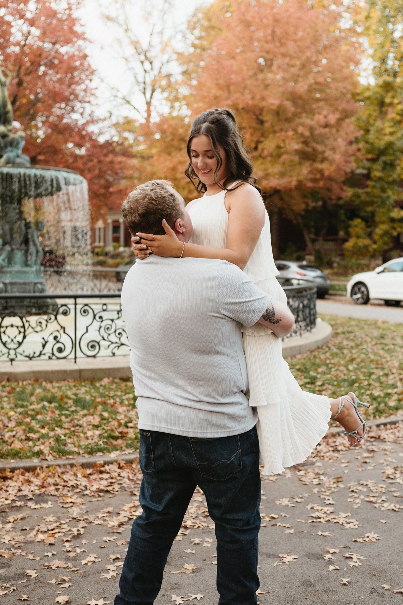 A couple shares playful moments on autumn sidewalk with orange fall foliage in the background.