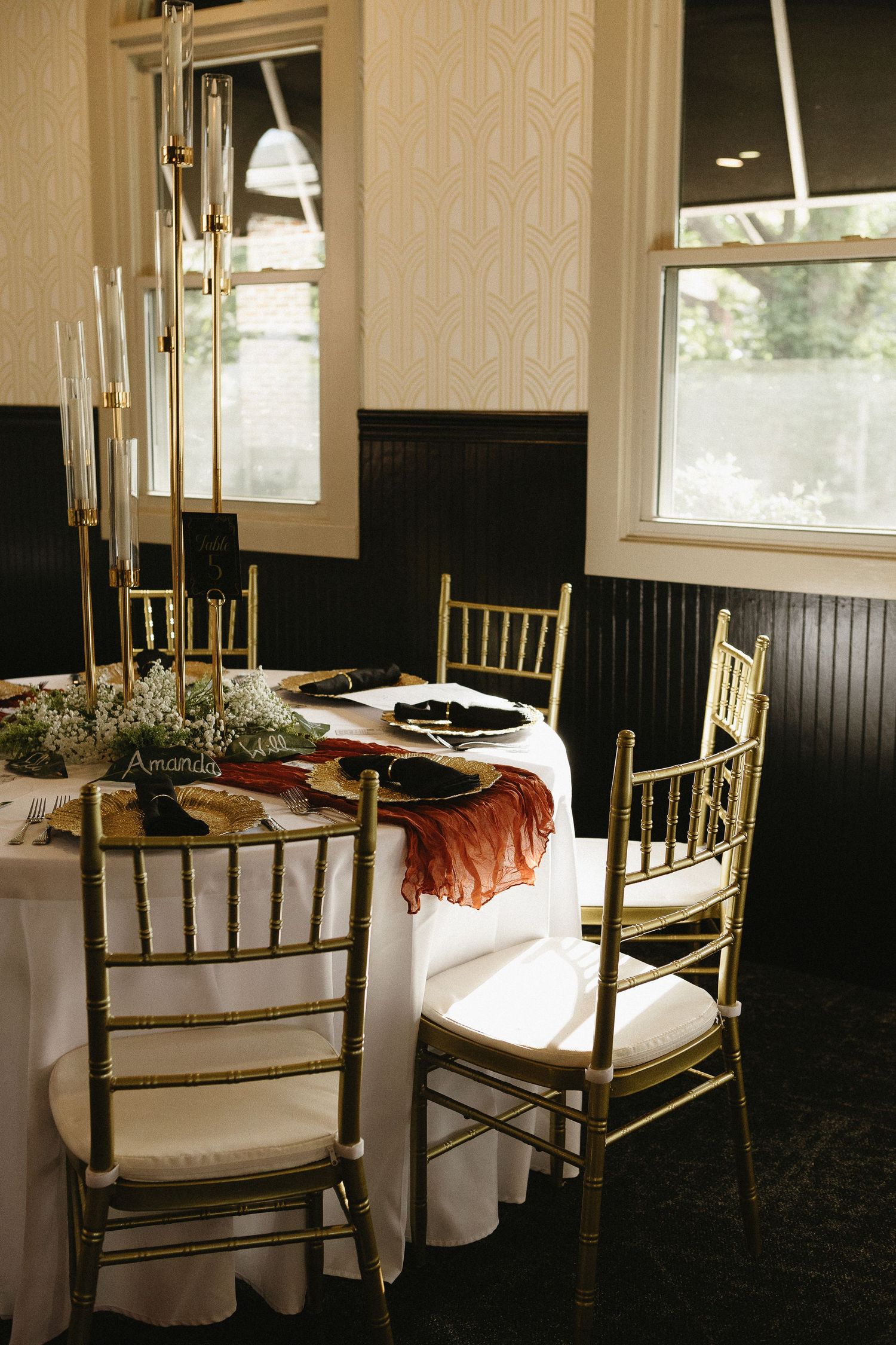 Elegant dining table setup with gold chiavari chairs and candlesticks in a room with black wainscoting.
