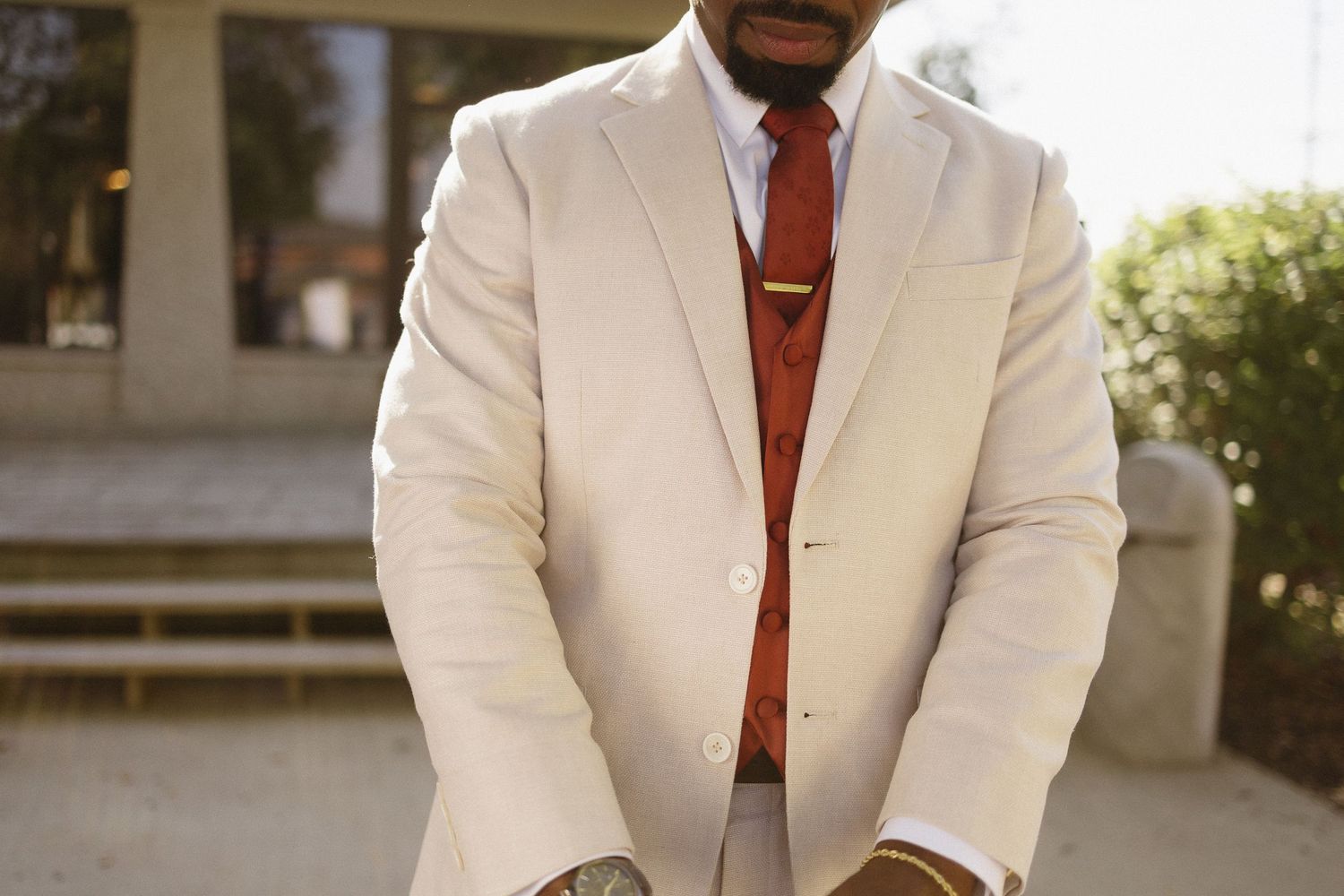 Groom in crisp white suit with burgundy tie poses outdoors.