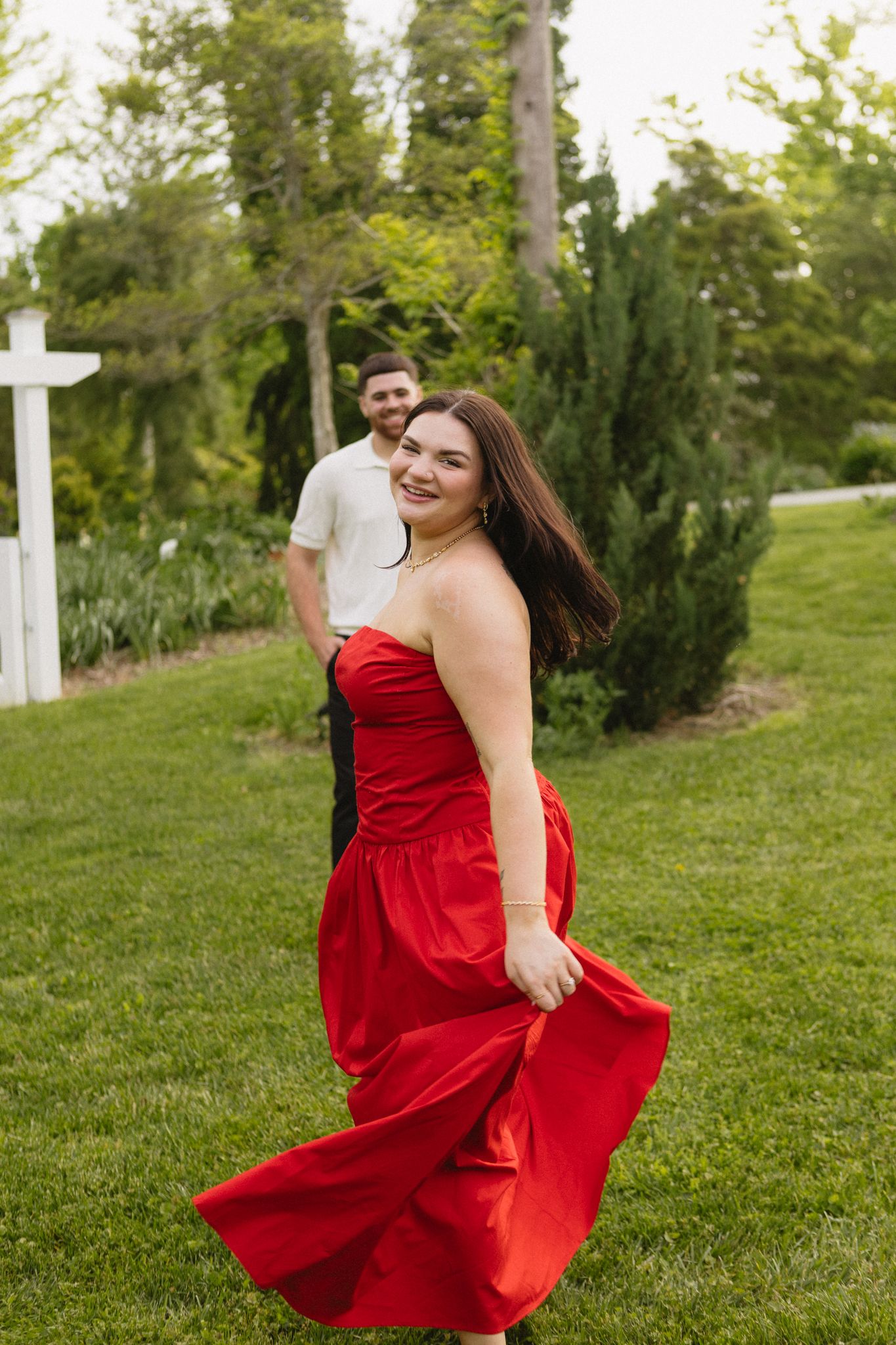 A person in a flowing red dress twirls on green grass near trees and white posts in a garden setting.