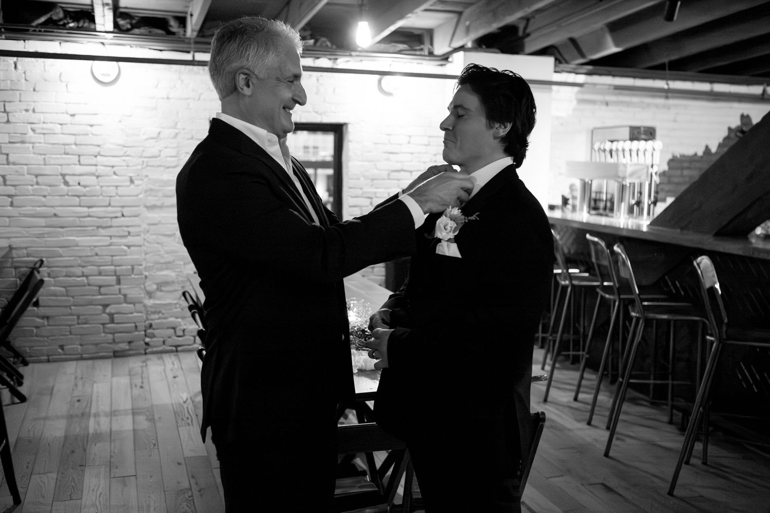 Two people in formal attire share a candid moment in a basement venue with exposed brick walls.
