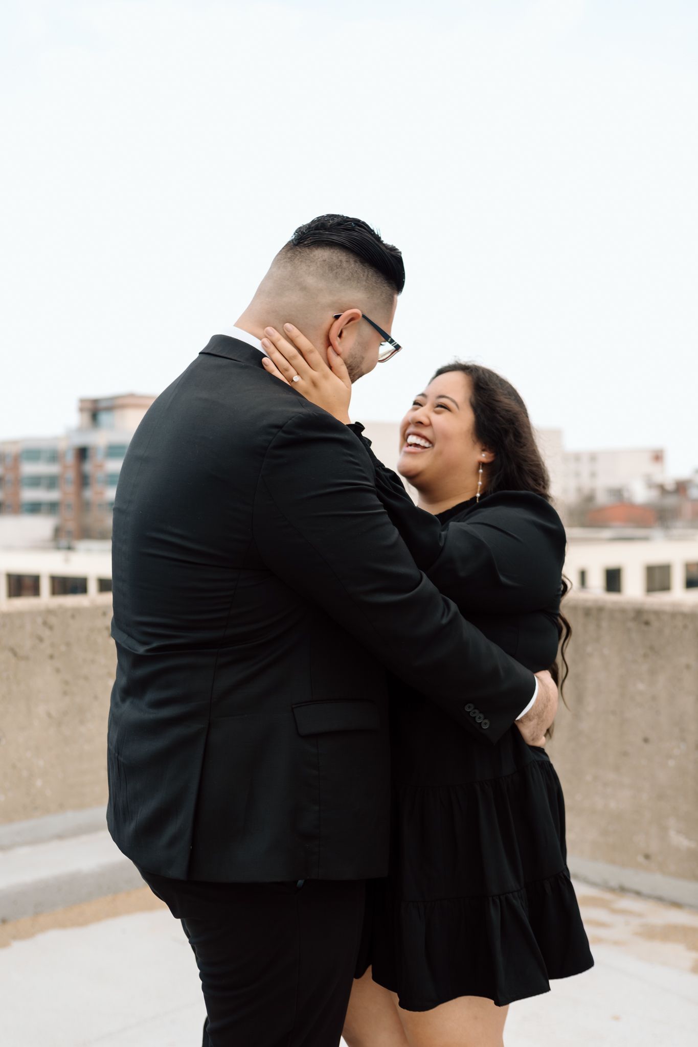 Elegant engaged couple smiling and embracing in downtown Louisville, with the woman showing her engagement ring