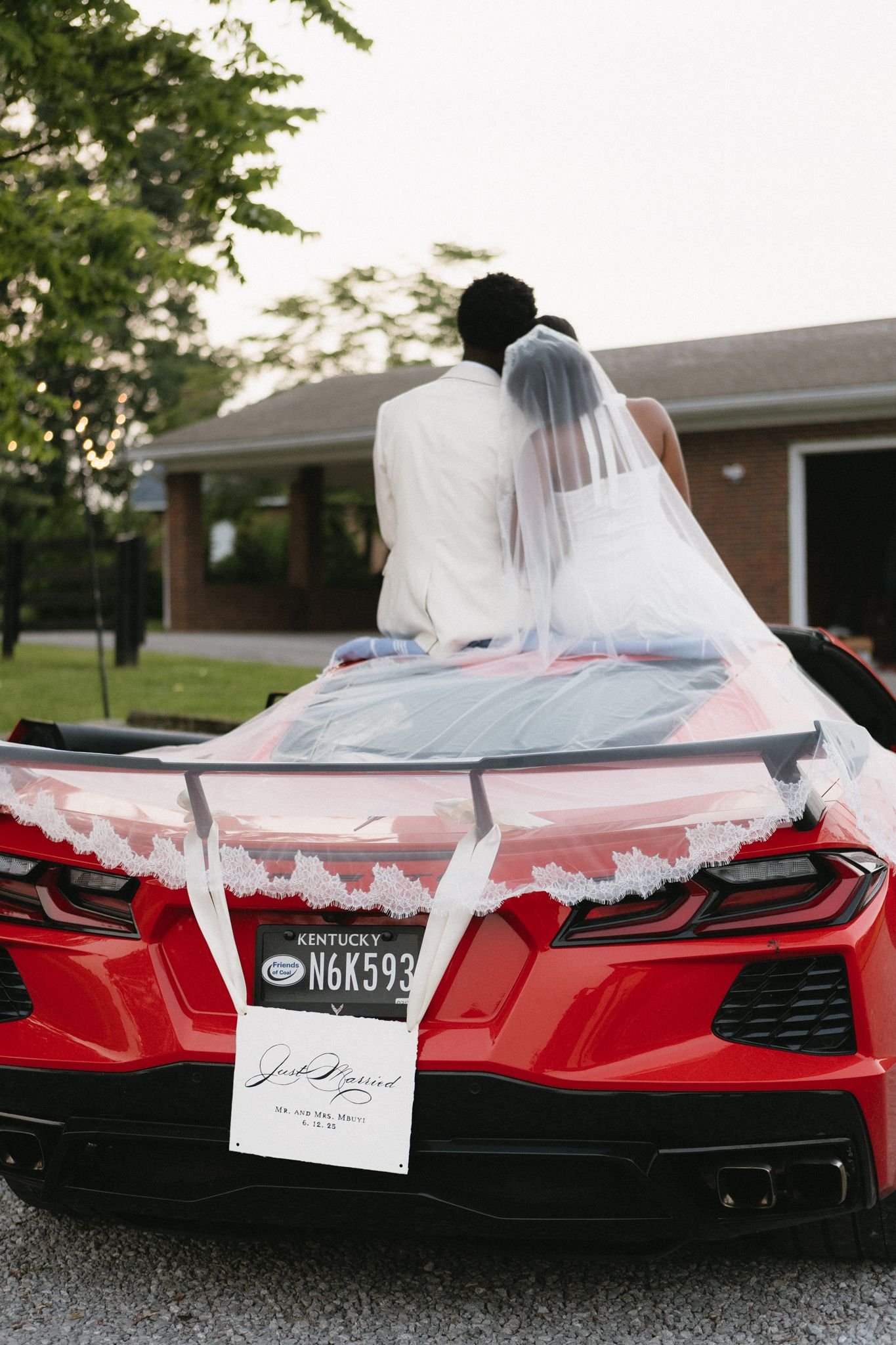 Newlyweds sit on a decorated red Corvette with a wedding sign and veil flowing.