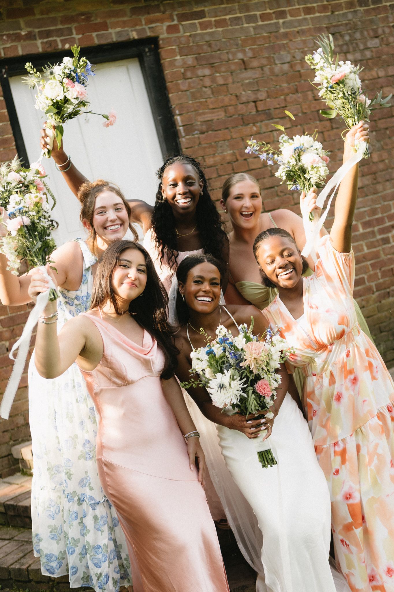 Enthusiastic bridal party celebrates against brick wall backdrop with floral bouquets.