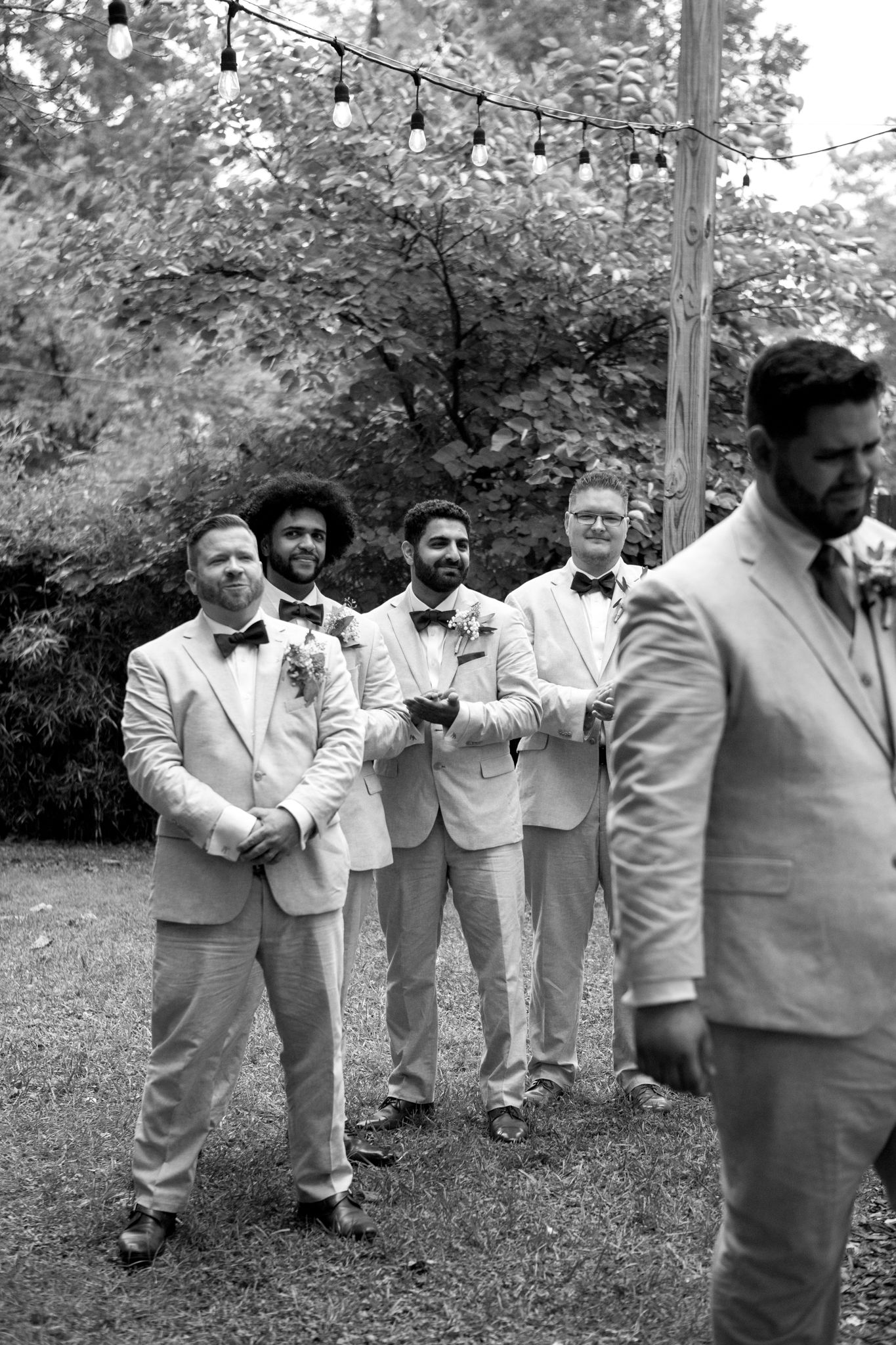 Groomsmen in light gray suits stand in line at an outdoor wedding with string lights overhead in black and white.
