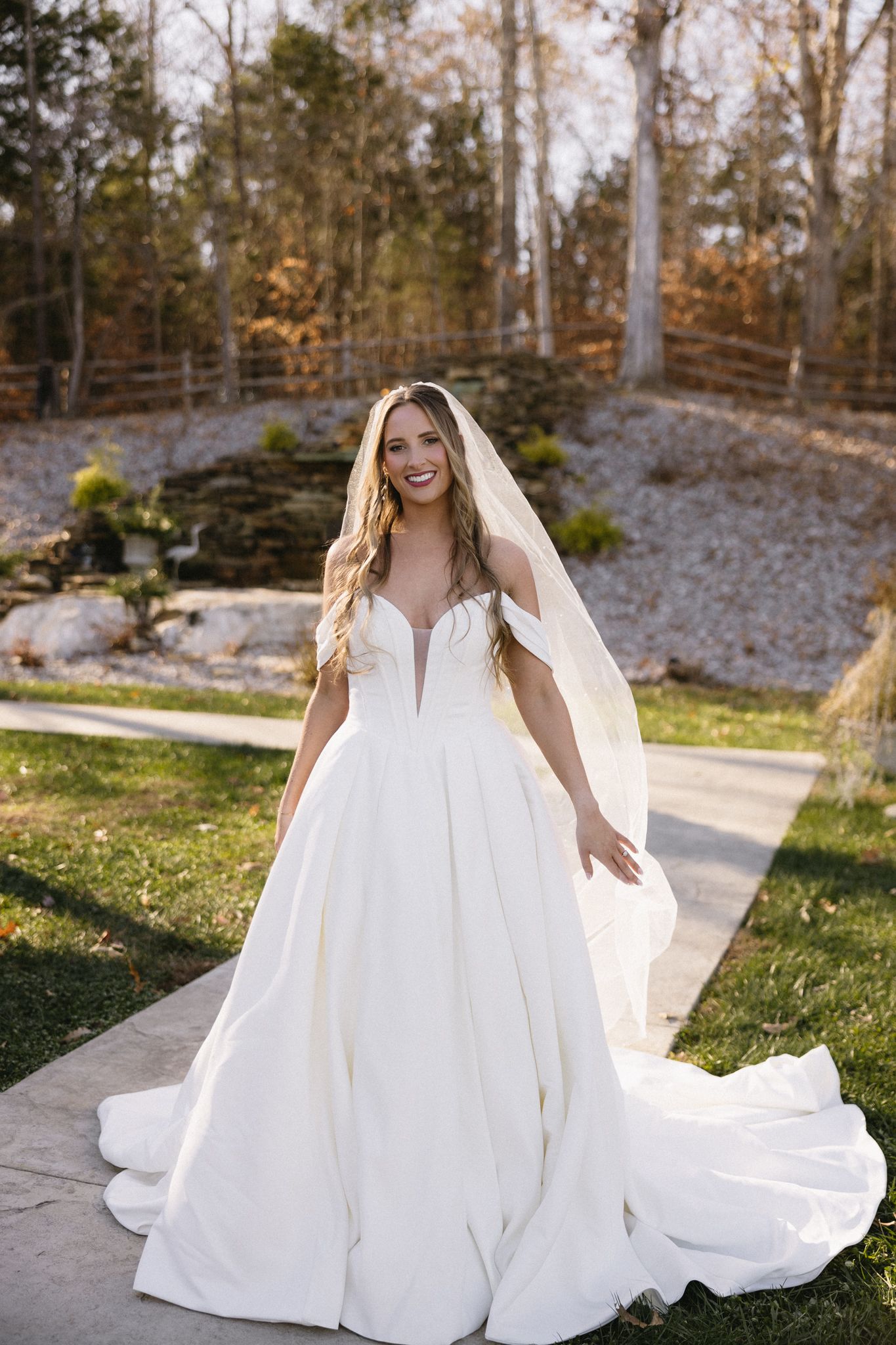 A bride in a flowing white ball gown and veil poses in front of an autumn forest backdrop.