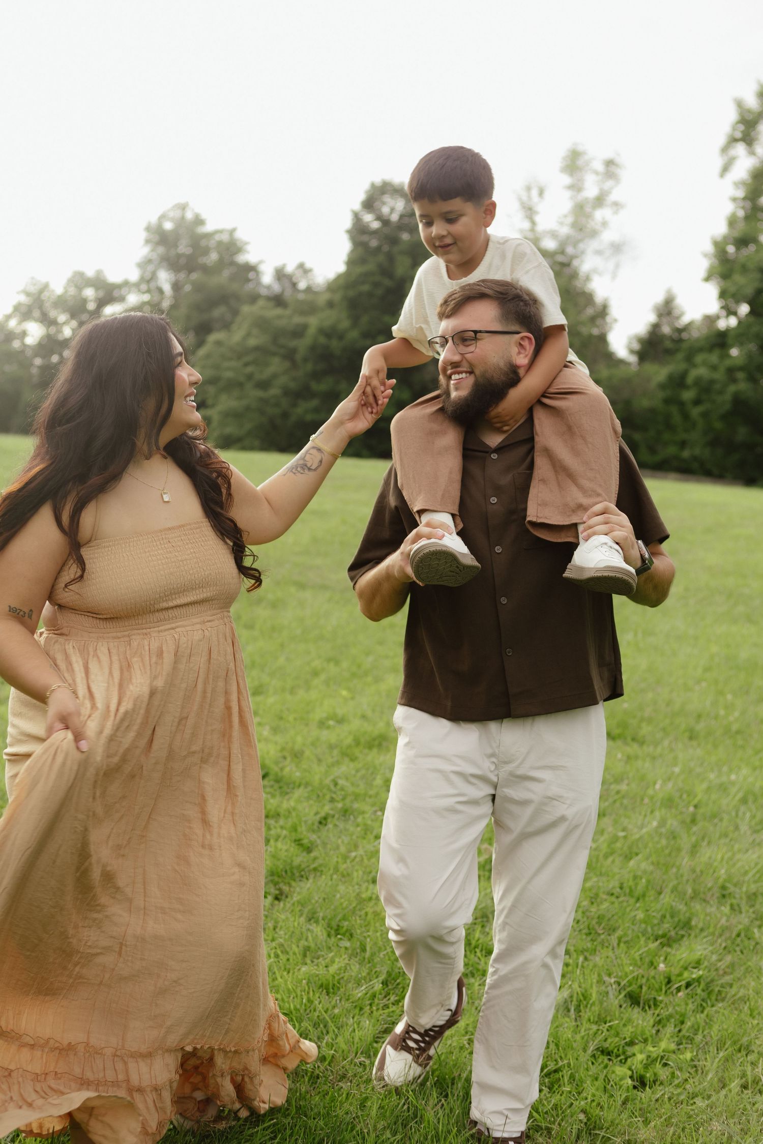 A happy family enjoys a sunny day outdoors, with dad carrying son on shoulders while mom smiles and holds hands.
