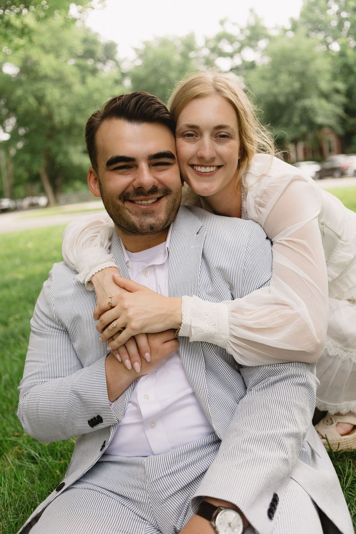 A smiling couple sits on grass in a park, the woman in white hugging the man in a gray suit from behind.