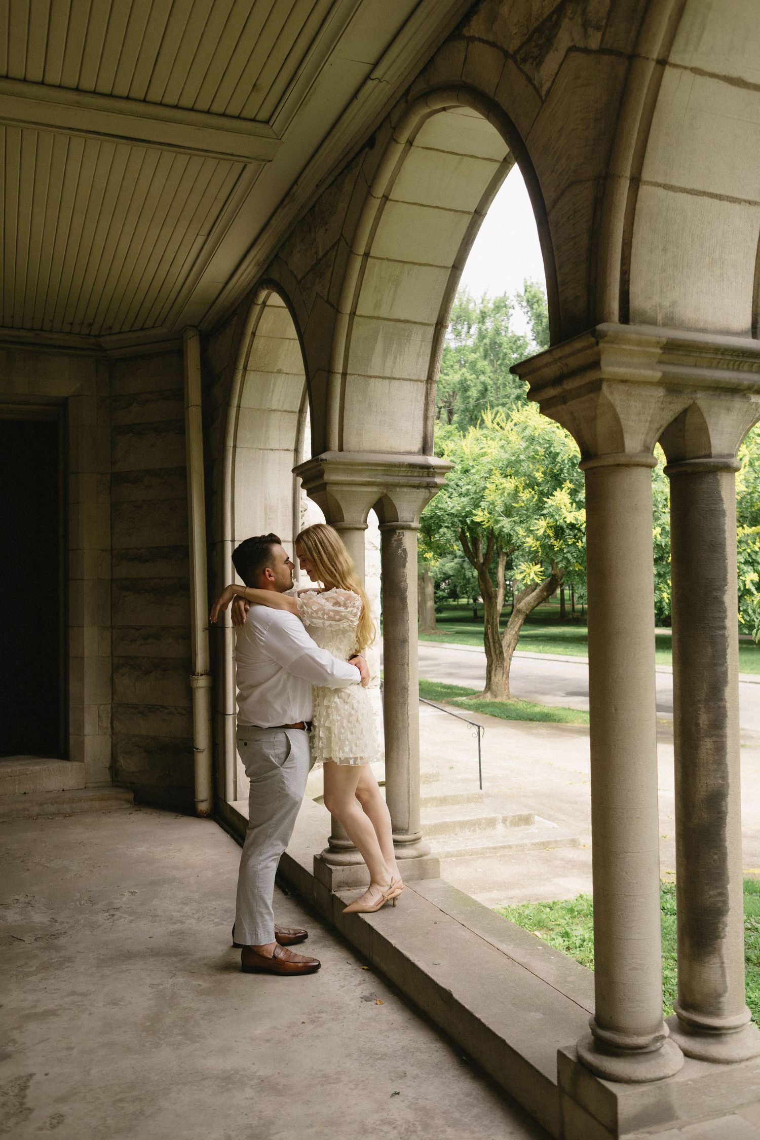 Couple embracing under stone arched colonnade in a lush garden setting.