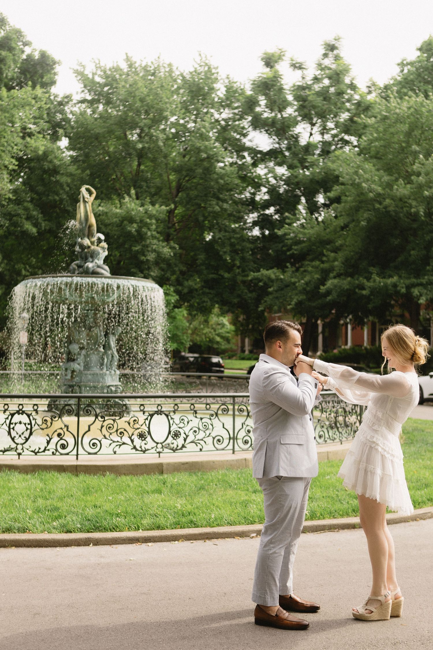 Couple in elegant attire poses by ornate fountain in lush park, woman adjusting man's tie outdoors.