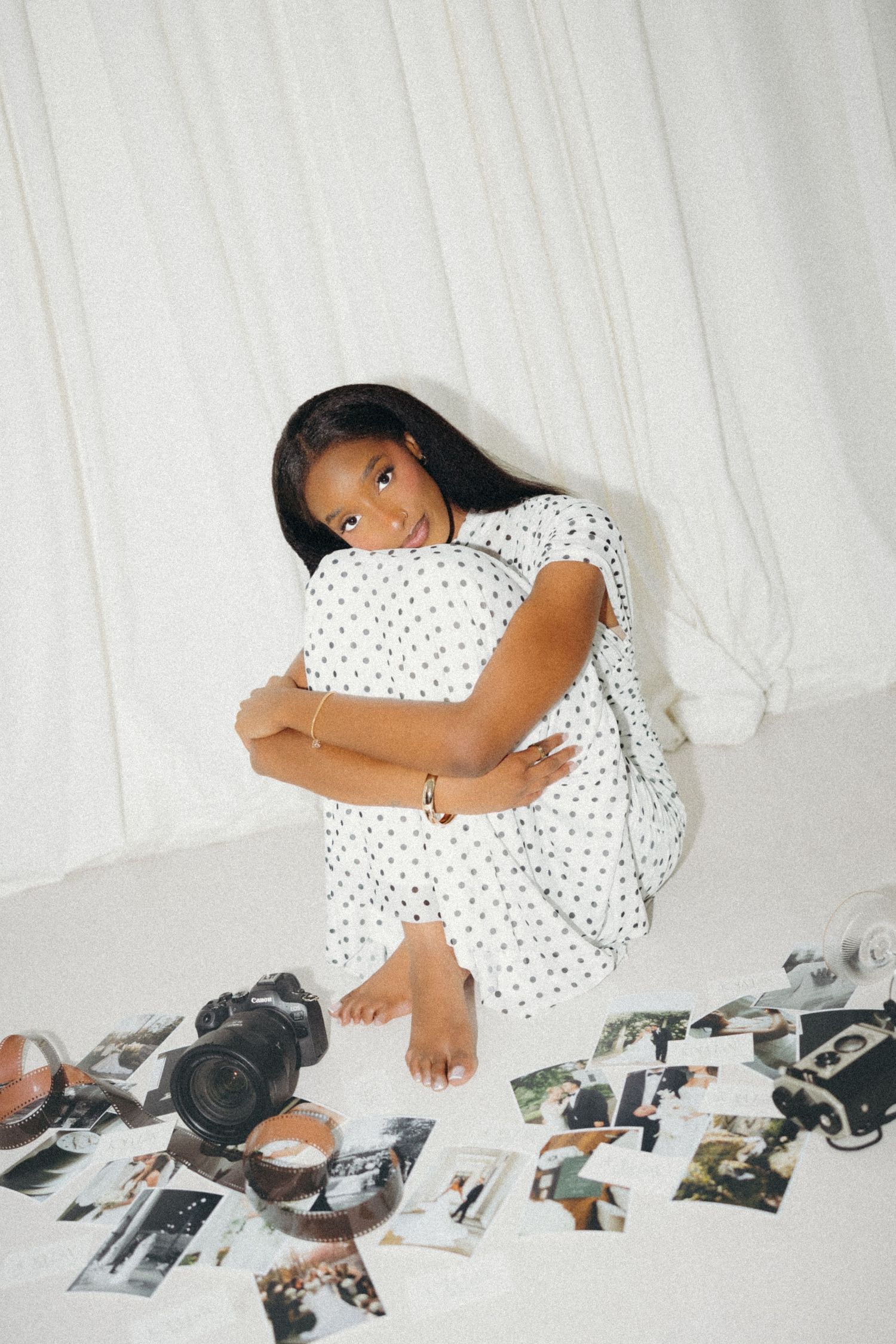 Young woman in white polka dot dress sitting on floor surrounded by photographs and cameras, hugging her knees.