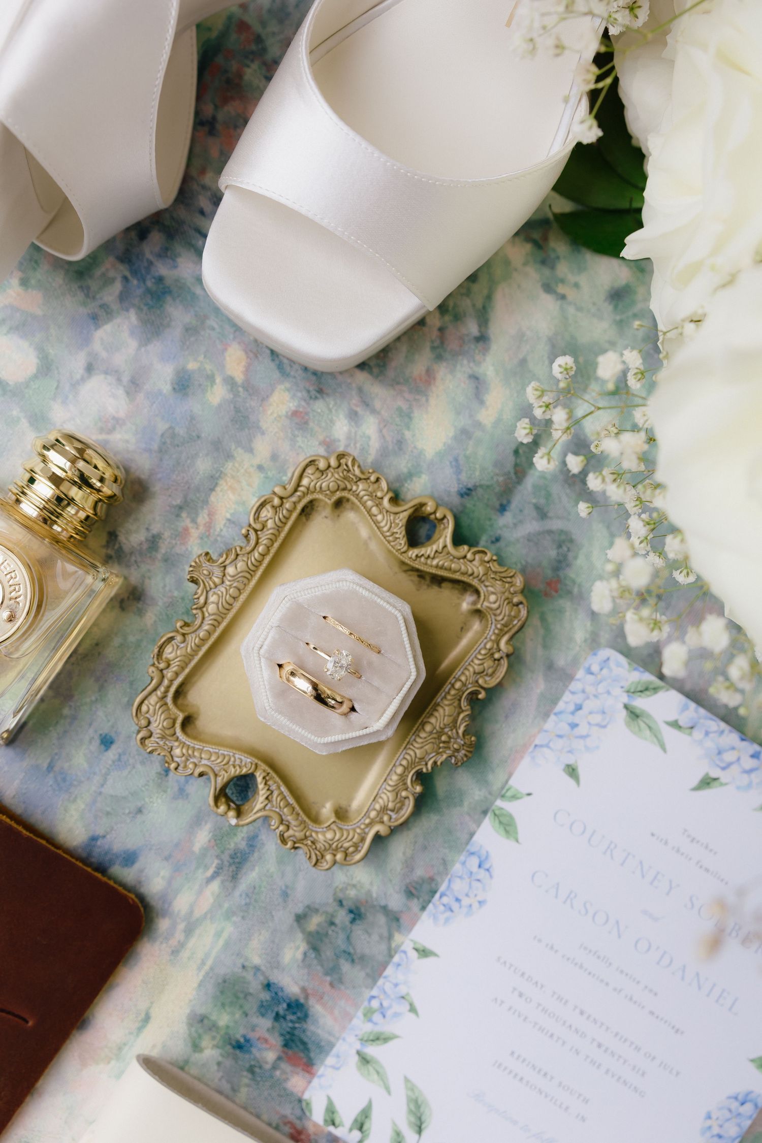 Elegant wedding flat lay with gold ring box, diamond rings, white heels, perfume, and floral invitation on watercolor surface.
