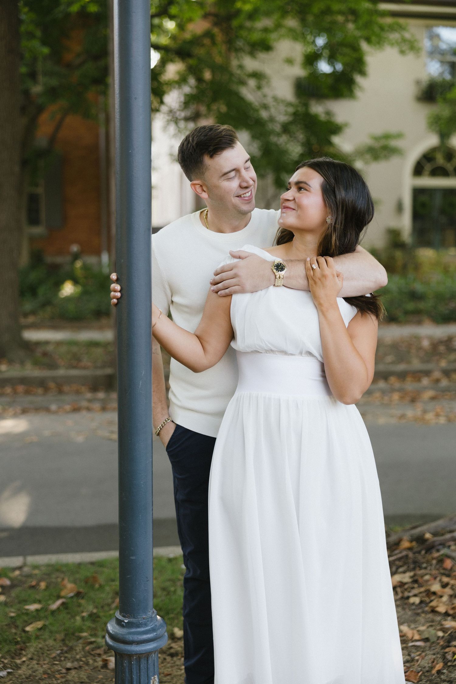 A couple embraces by a lamppost outdoors, woman in white dress, man in cream sweater, smiling at each other.