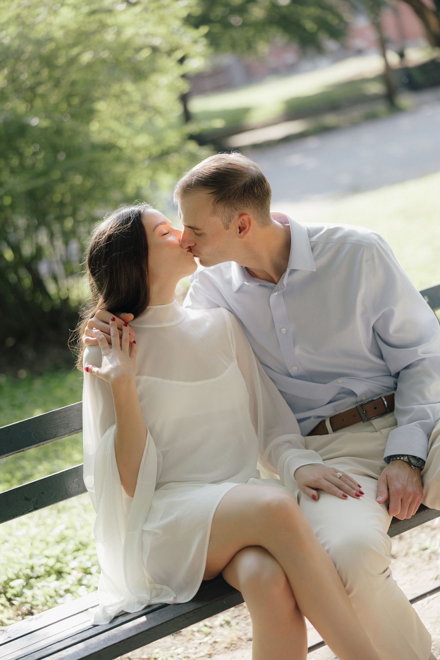 Romantic couple sharing a kiss on a park bench surrounded by lush green trees.