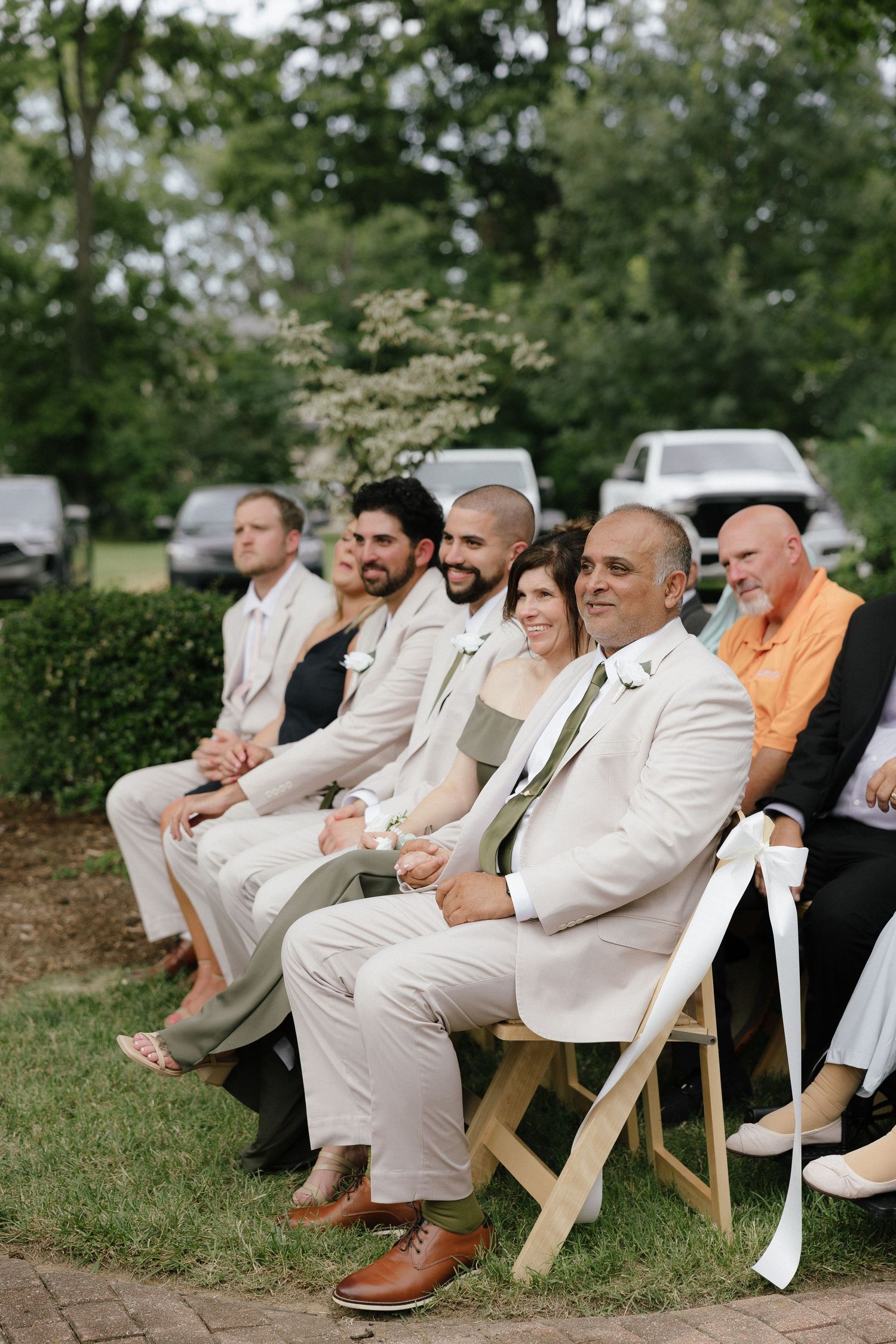 Wedding guests in tan suits and casual attire seated in white folding chairs at outdoor ceremony.