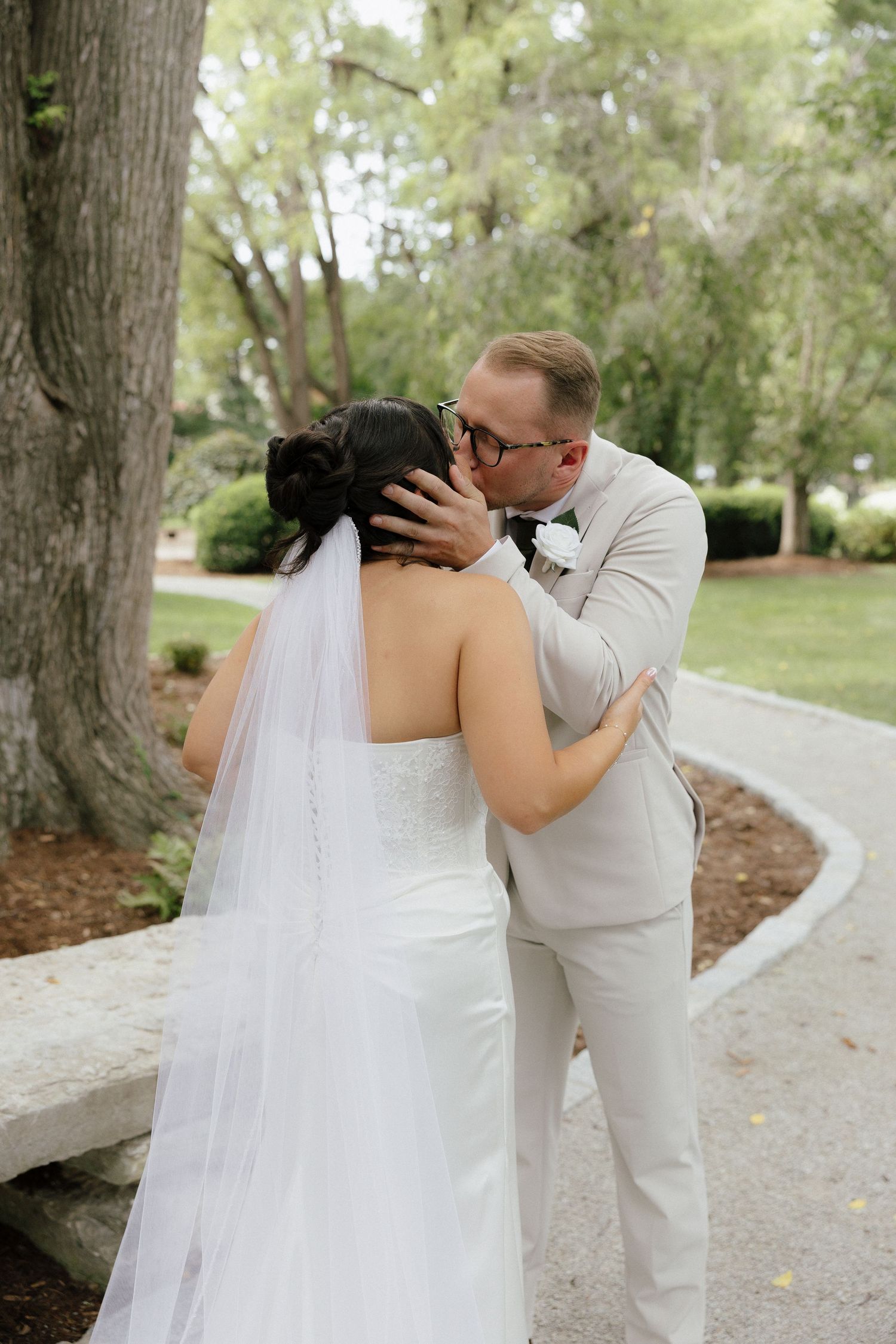 Groom in light gray suit tenderly kissing bride in white gown and veil on garden pathway.