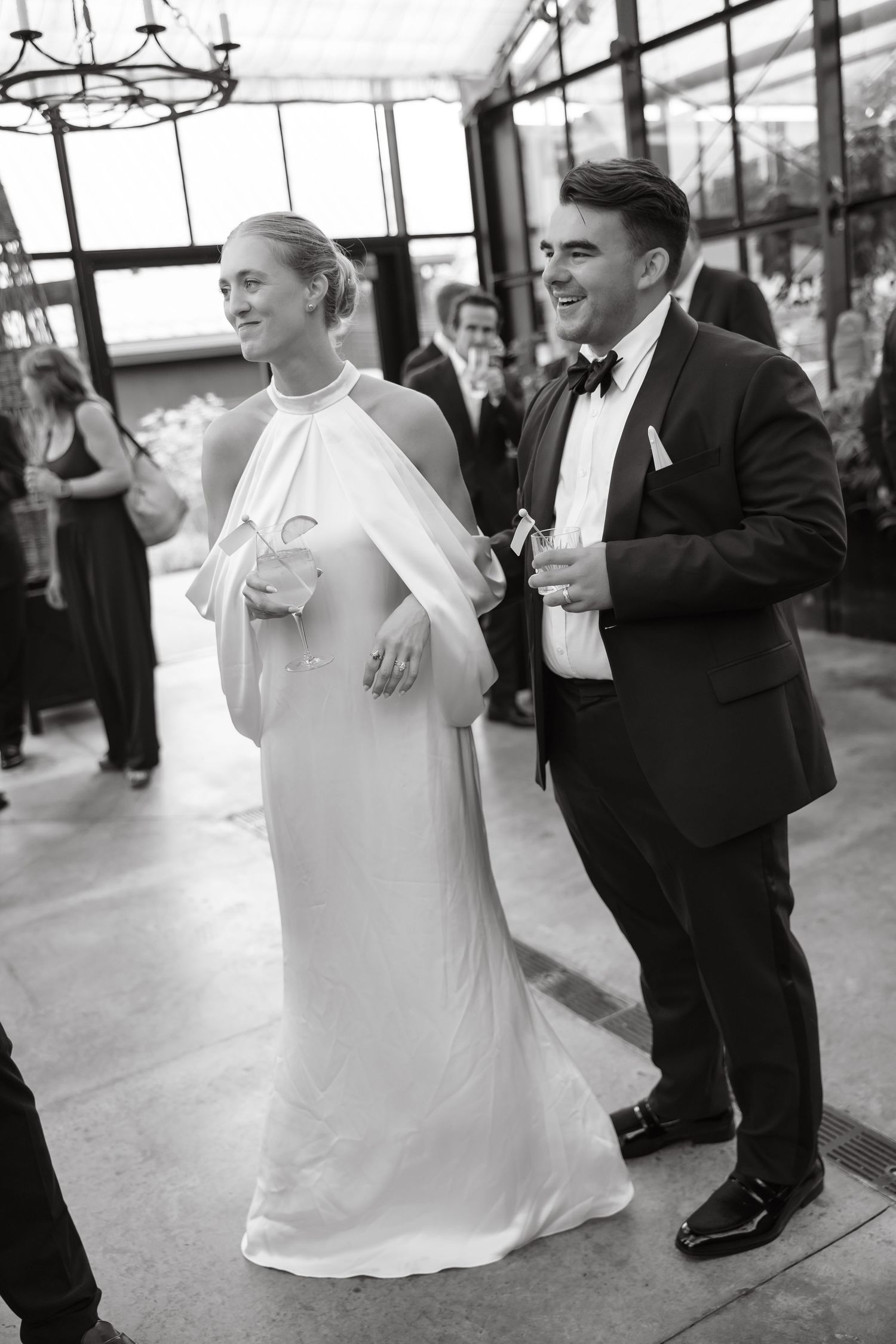 Bride in flowing white halter gown and groom in tuxedo laugh together at indoor wedding reception.