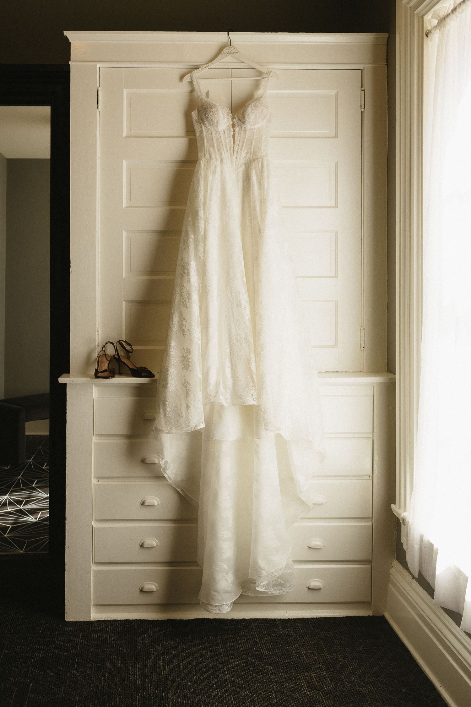 A white wedding dress hangs elegantly against a white closet door in natural light.