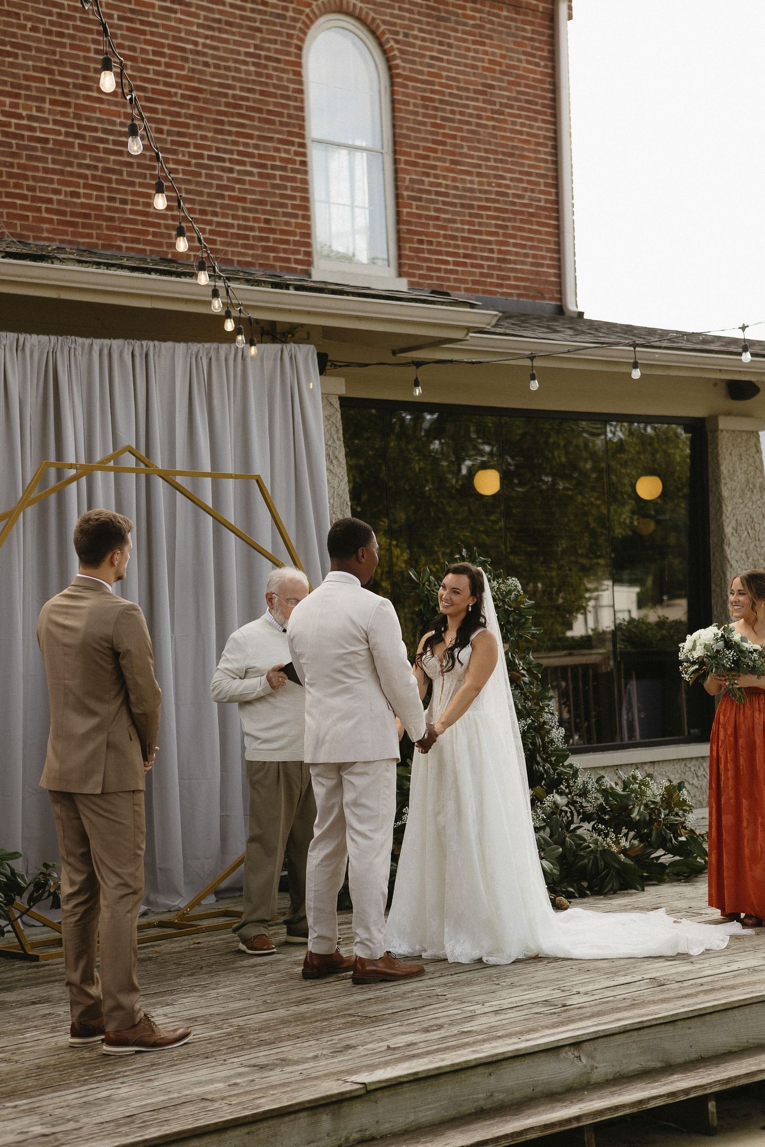 Wedding ceremony taking place on a wooden deck with string lights and brick backdrop.