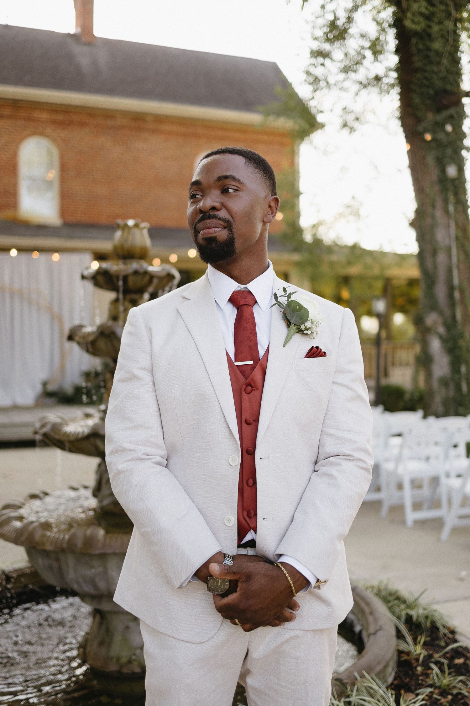 Groom in white suit with red tie standing in an outdoor wedding venue.