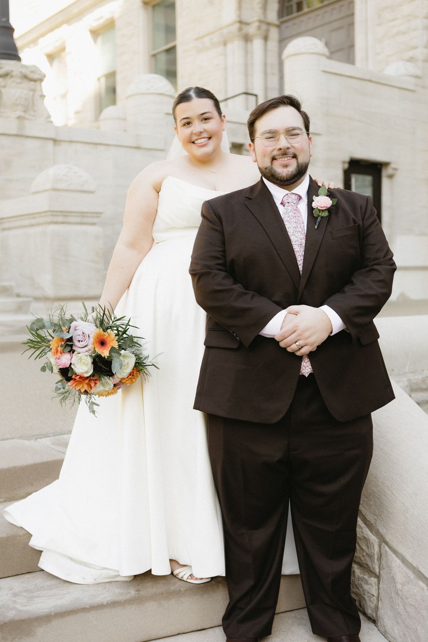 A bride and groom pose together on stone steps outside a classic building with columns.