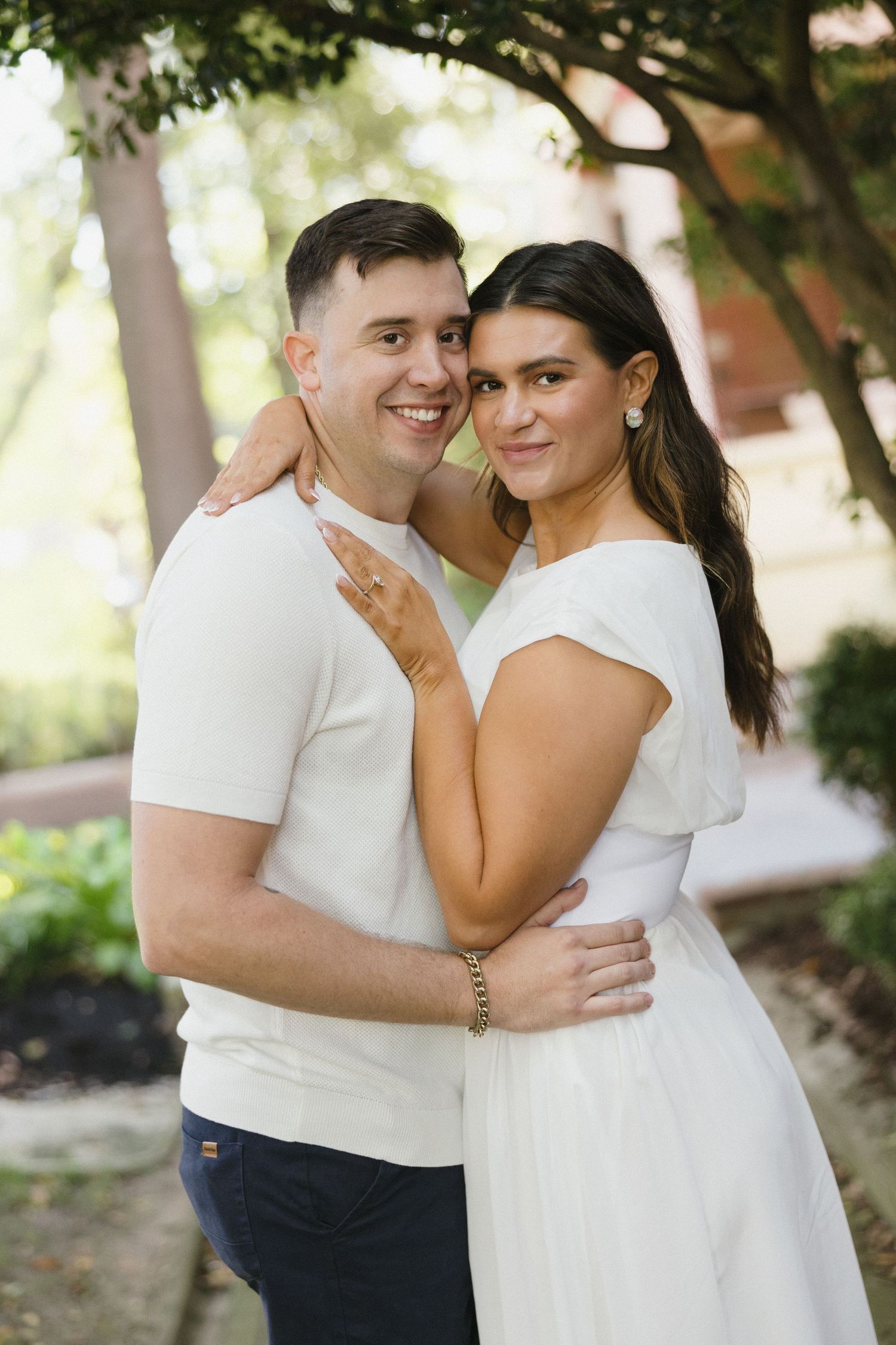 Engaged couple embracing outdoors under trees, woman in white dress and man in white shirt smiling.