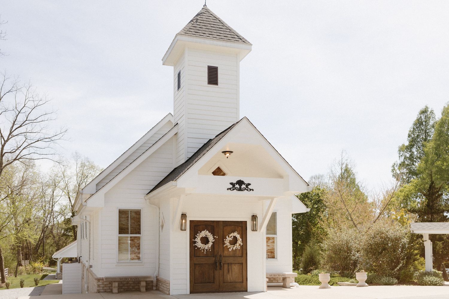 A charming white country chapel with a bell tower and rustic wooden doors decorated with a seasonal wreath.