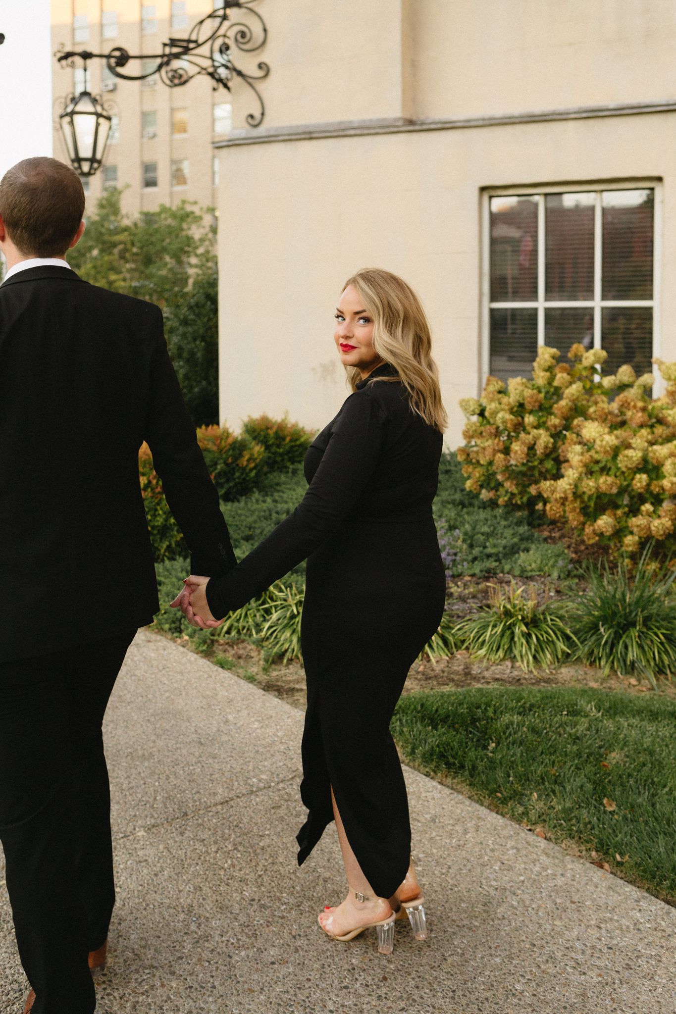 Couple in formal black attire holding hands while walking on garden path near yellow flowers and ornate wall lantern.