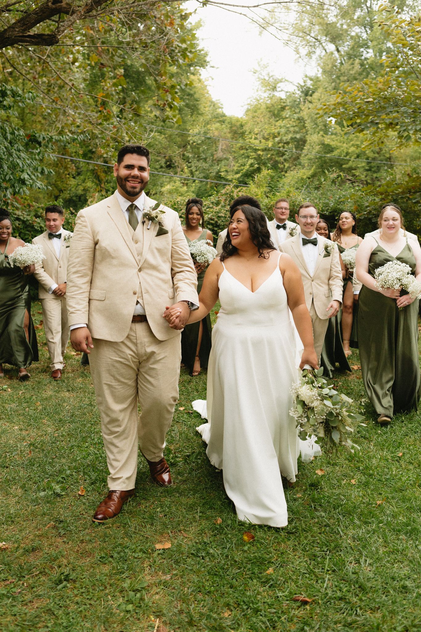 Joyful wedding party in beige suits and green dresses poses together on grassy lawn surrounded by trees.