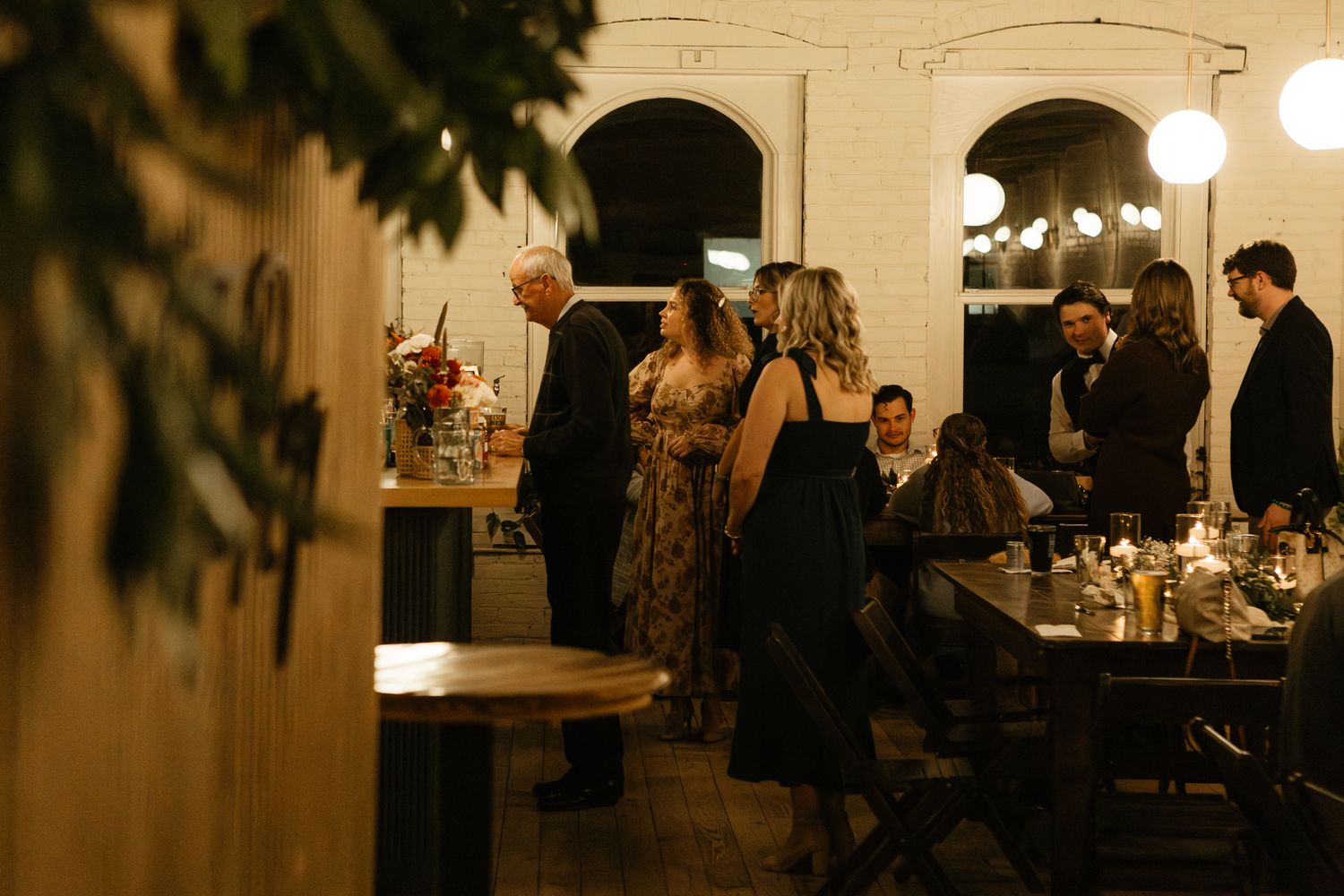 Wedding reception guests gather around tables in a warmly lit venue with brick walls.