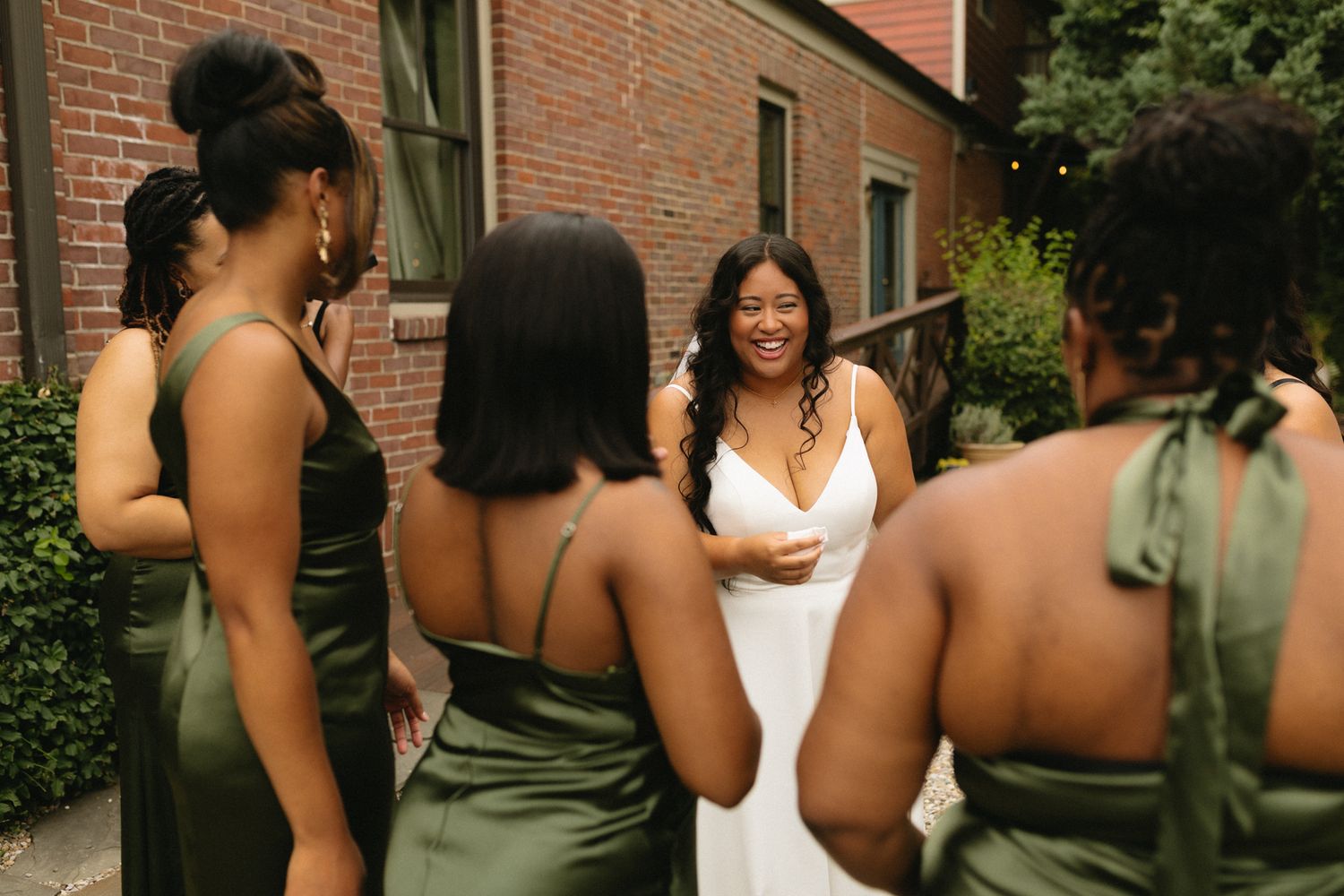 A group in olive green bridesmaid dresses gather around a person wearing white during a wedding celebration.
