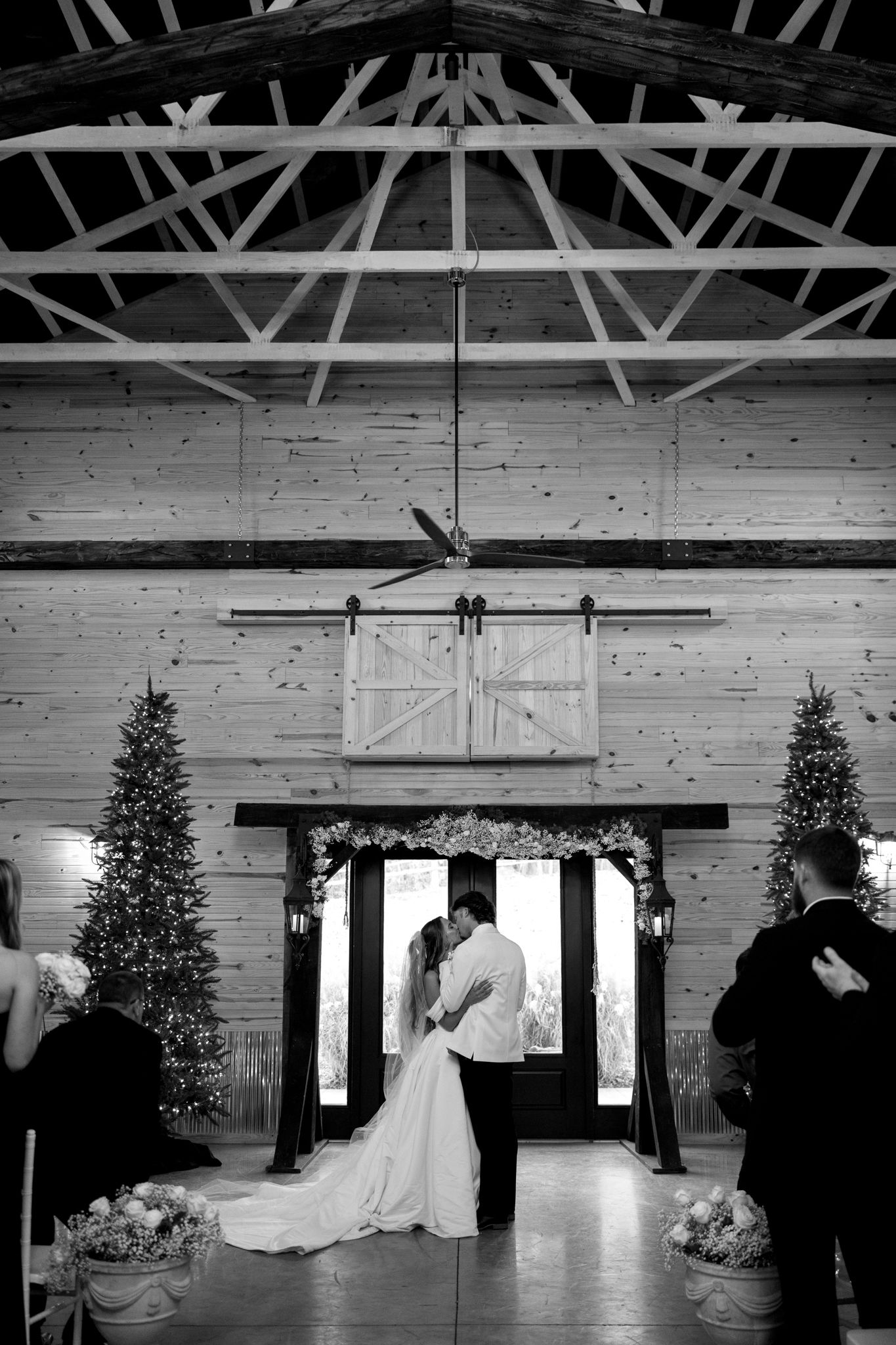 A rustic wedding venue interior with exposed wooden beams, white walls, and Christmas trees along the windows.