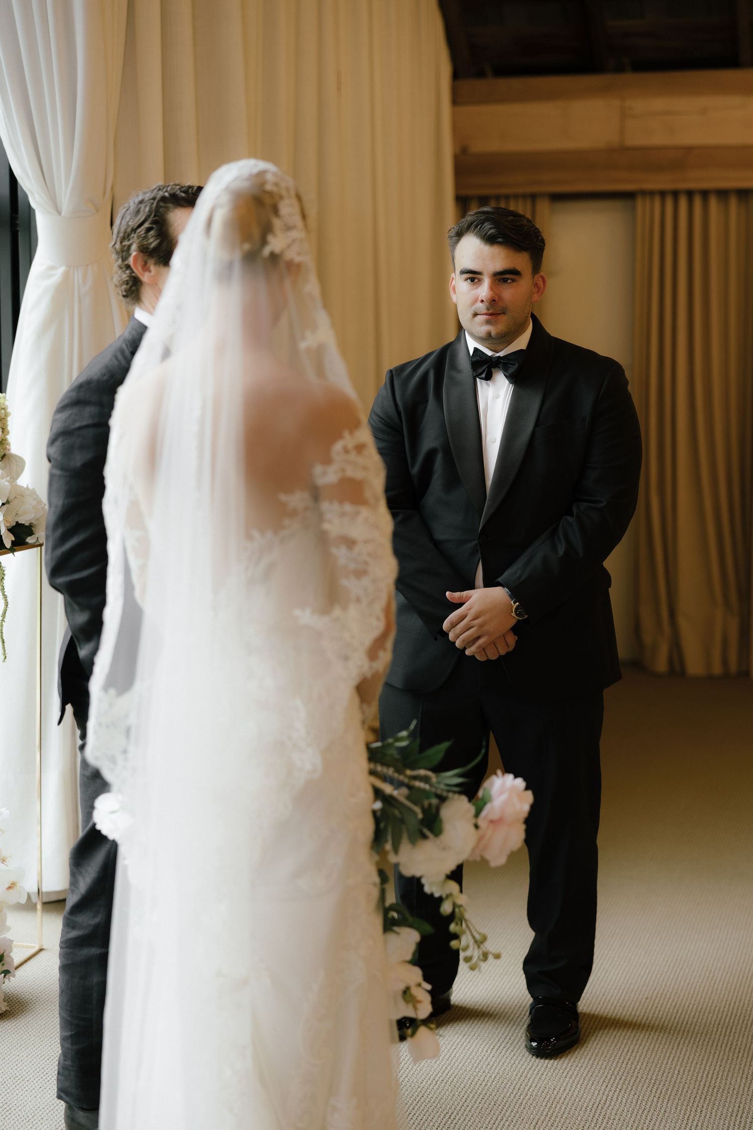Groom in black tuxedo watches bride approach during indoor wedding ceremony with white draping.