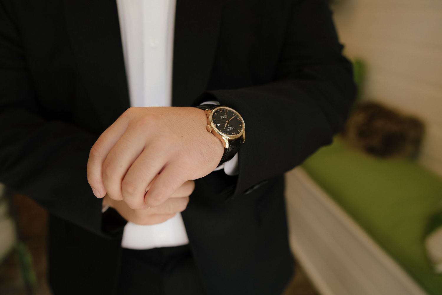 Groom in black suit adjusting gold watch on wrist before wedding ceremony.