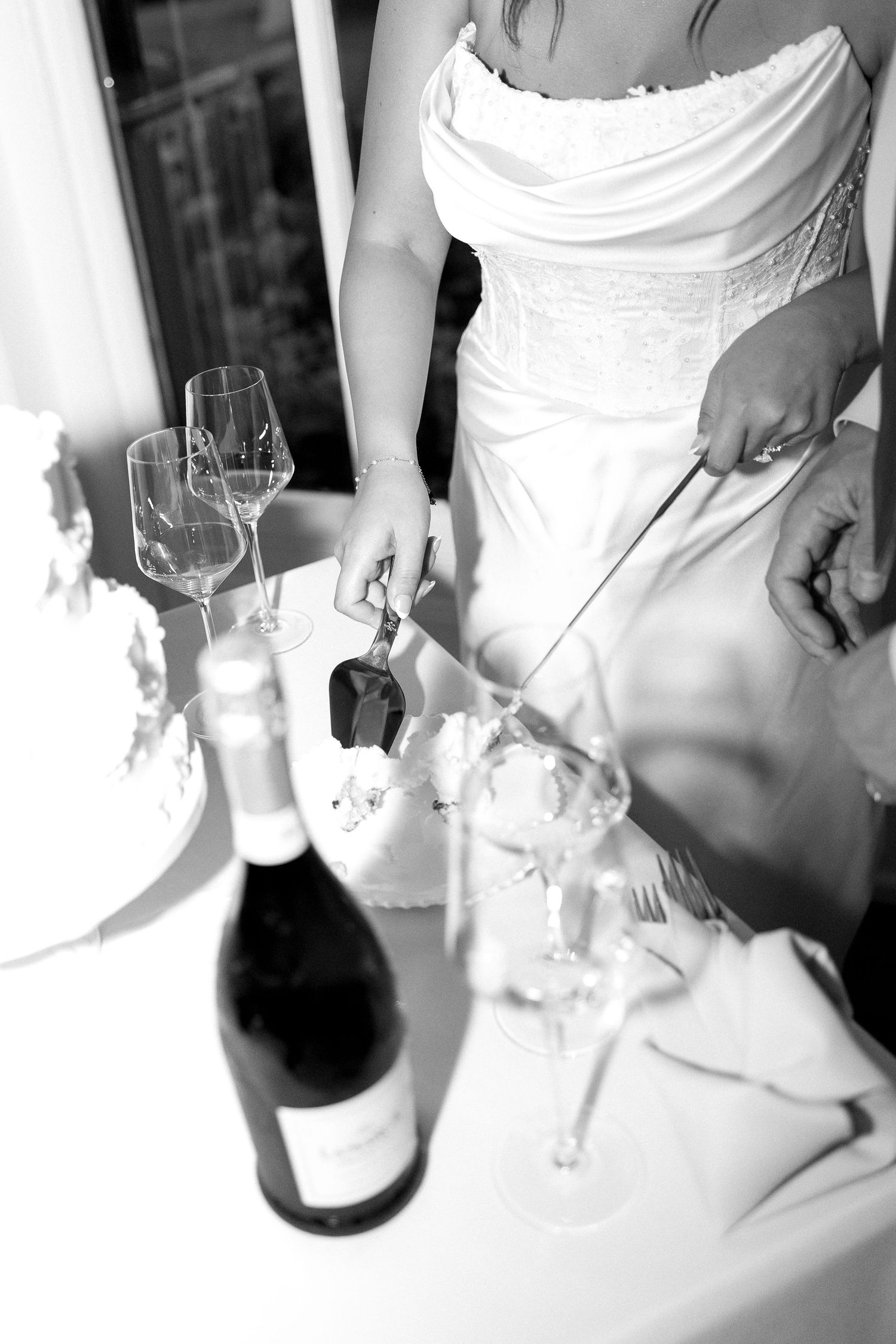 Black and white photo of bride cutting wedding cake beside champagne bottle and wine glasses.