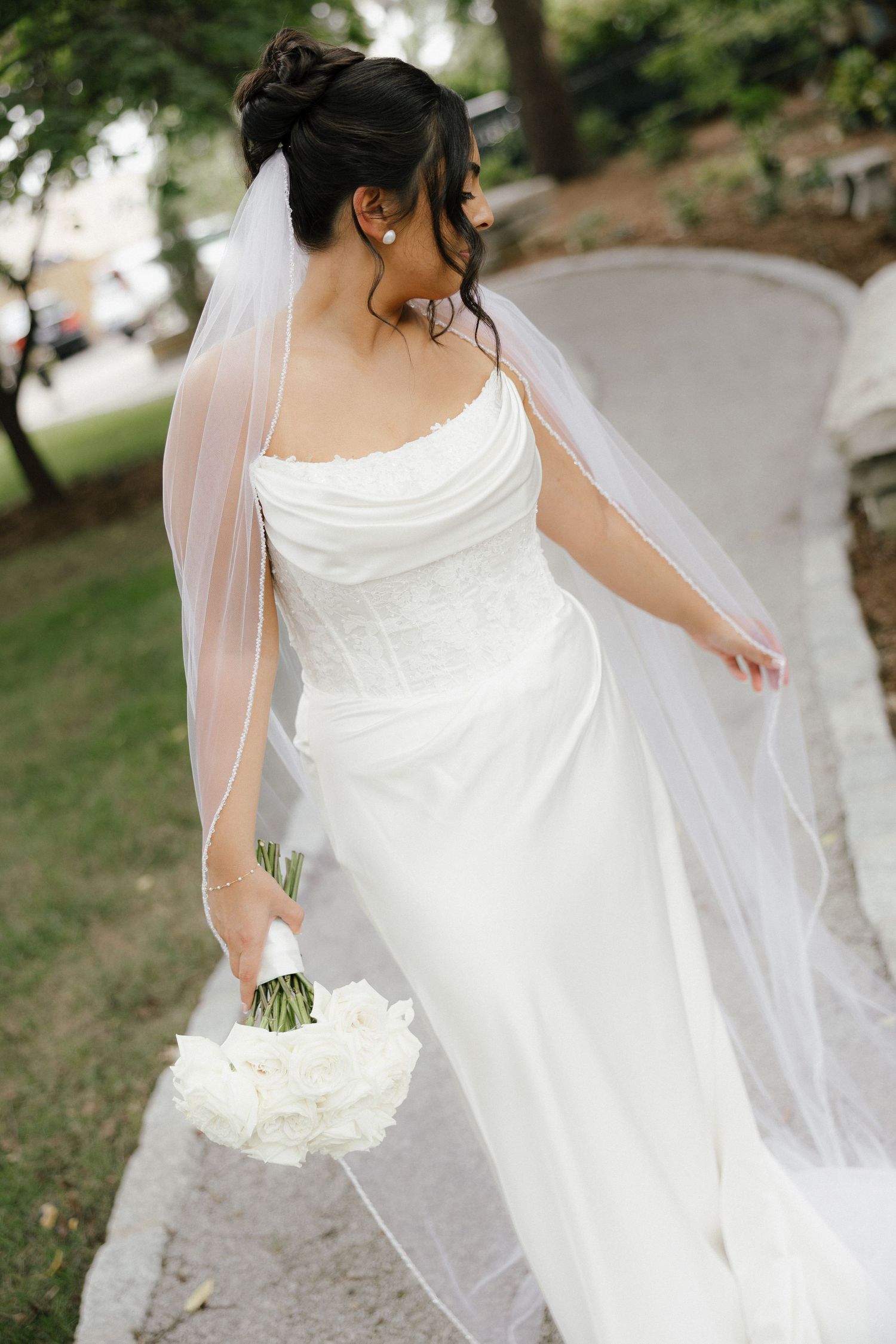 Bride in strapless white gown with flowing veil holds white bouquet while walking through outdoor garden.