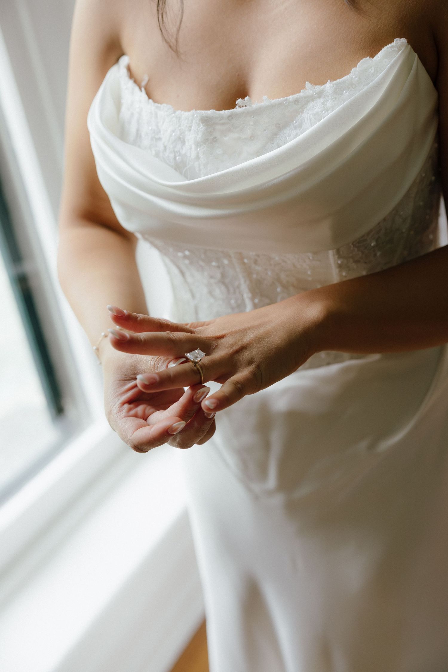 Bride in a white strapless wedding gown putting on her diamond engagement ring near a bright window.