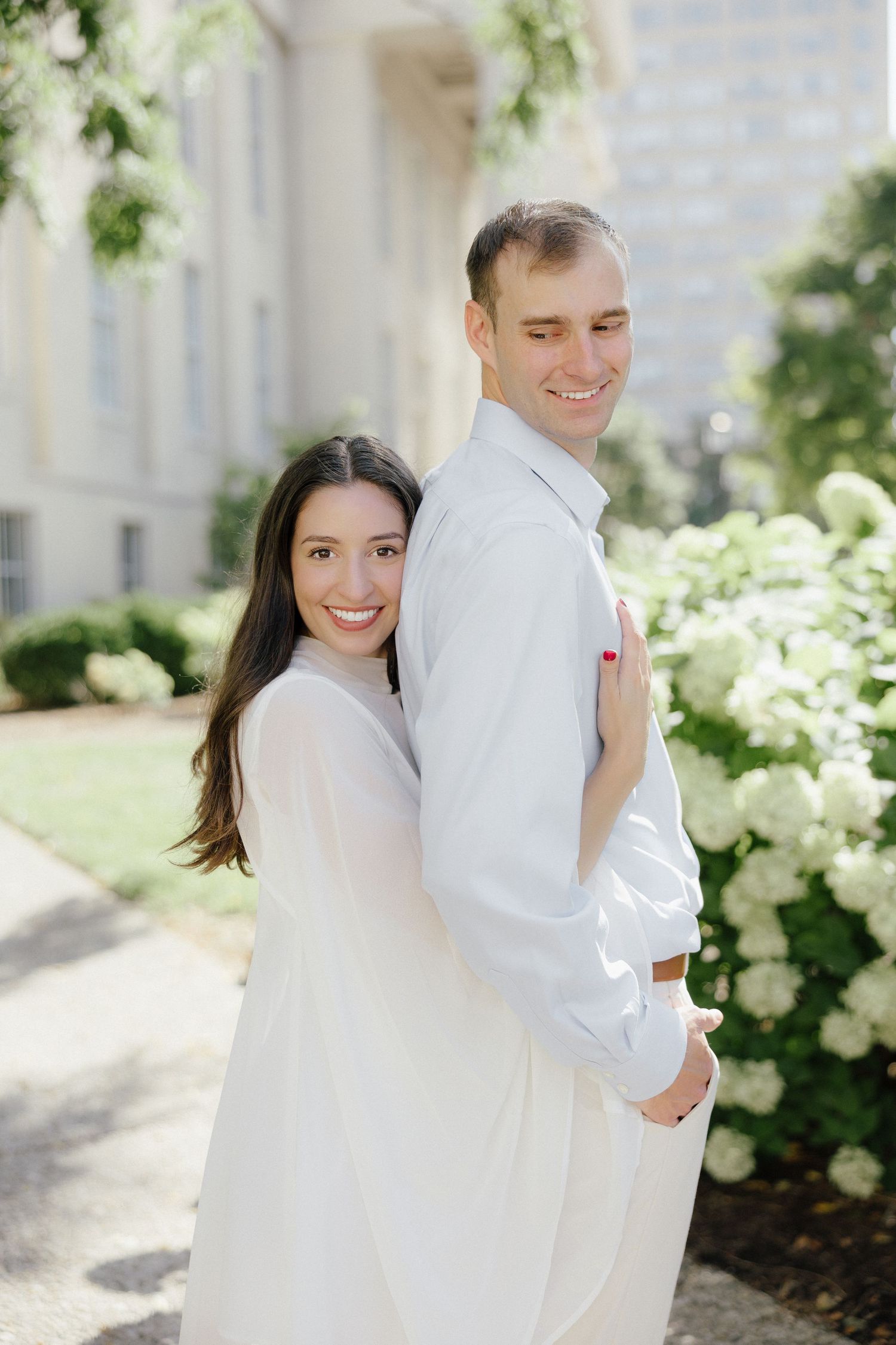 Happy couple embracing near blooming white hydrangeas outside a grand white building on a sunny day.